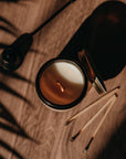 Candle in a glass holder on a wooden surface with a pen and leaf shadows.