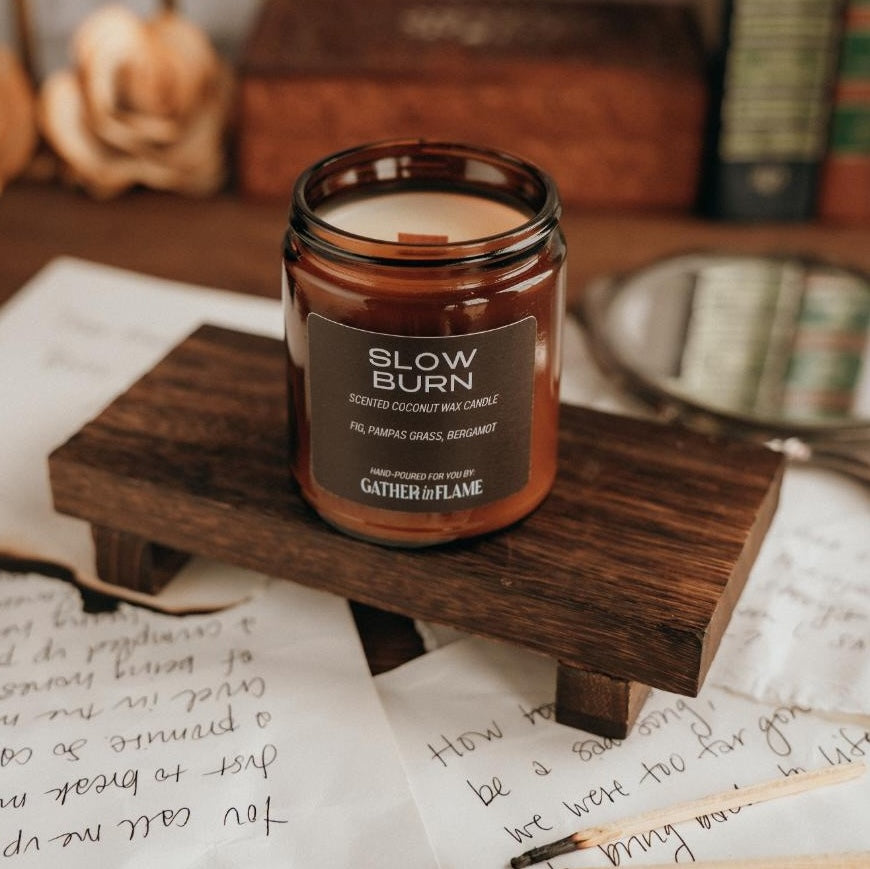 Amber glass jar candle labeled Slow Burn on a wood pedestal surrounded by love notes and matches