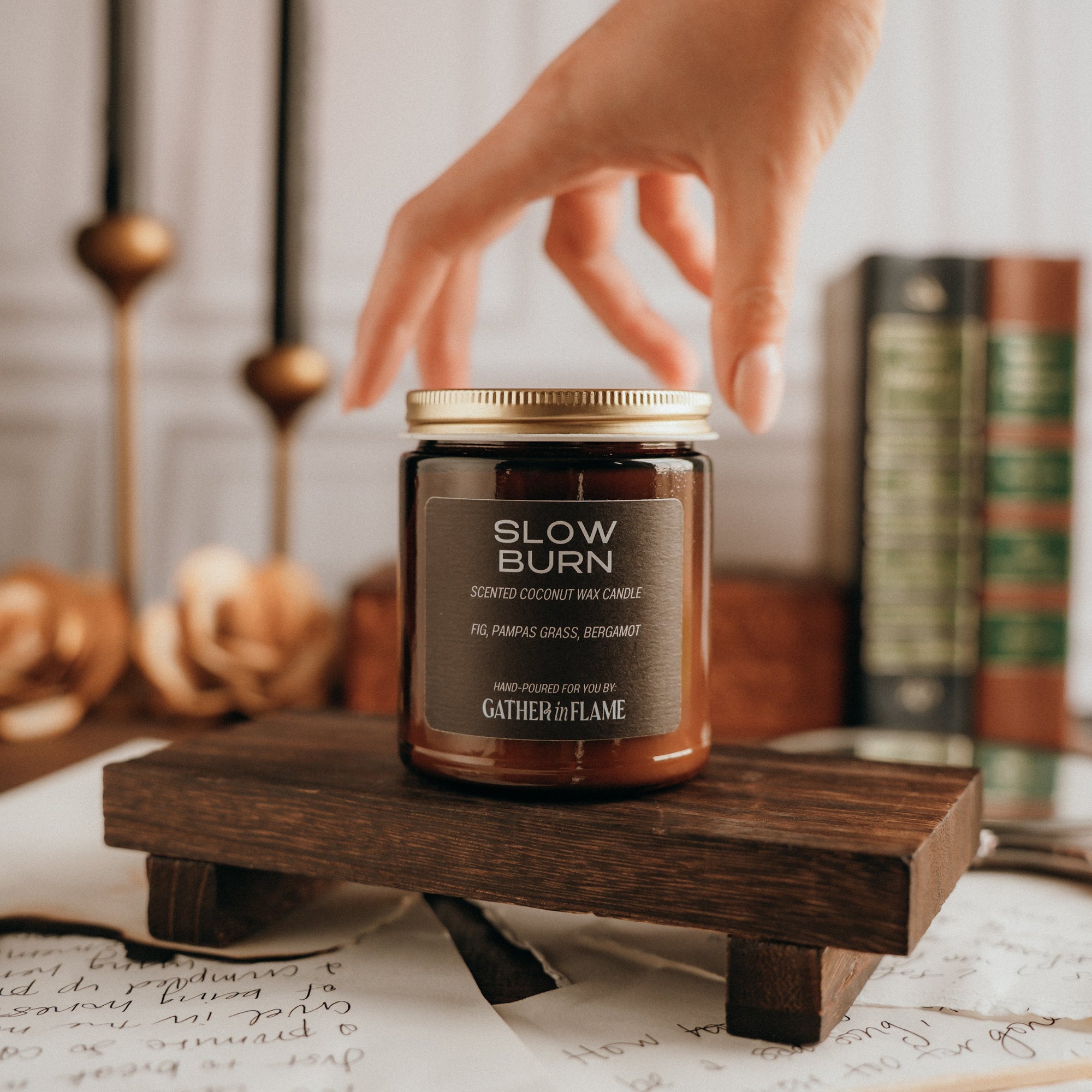 Jar of 'Slow Burn' candle on a wooden stand with a hand reaching towards it, surrounded by books and candles.