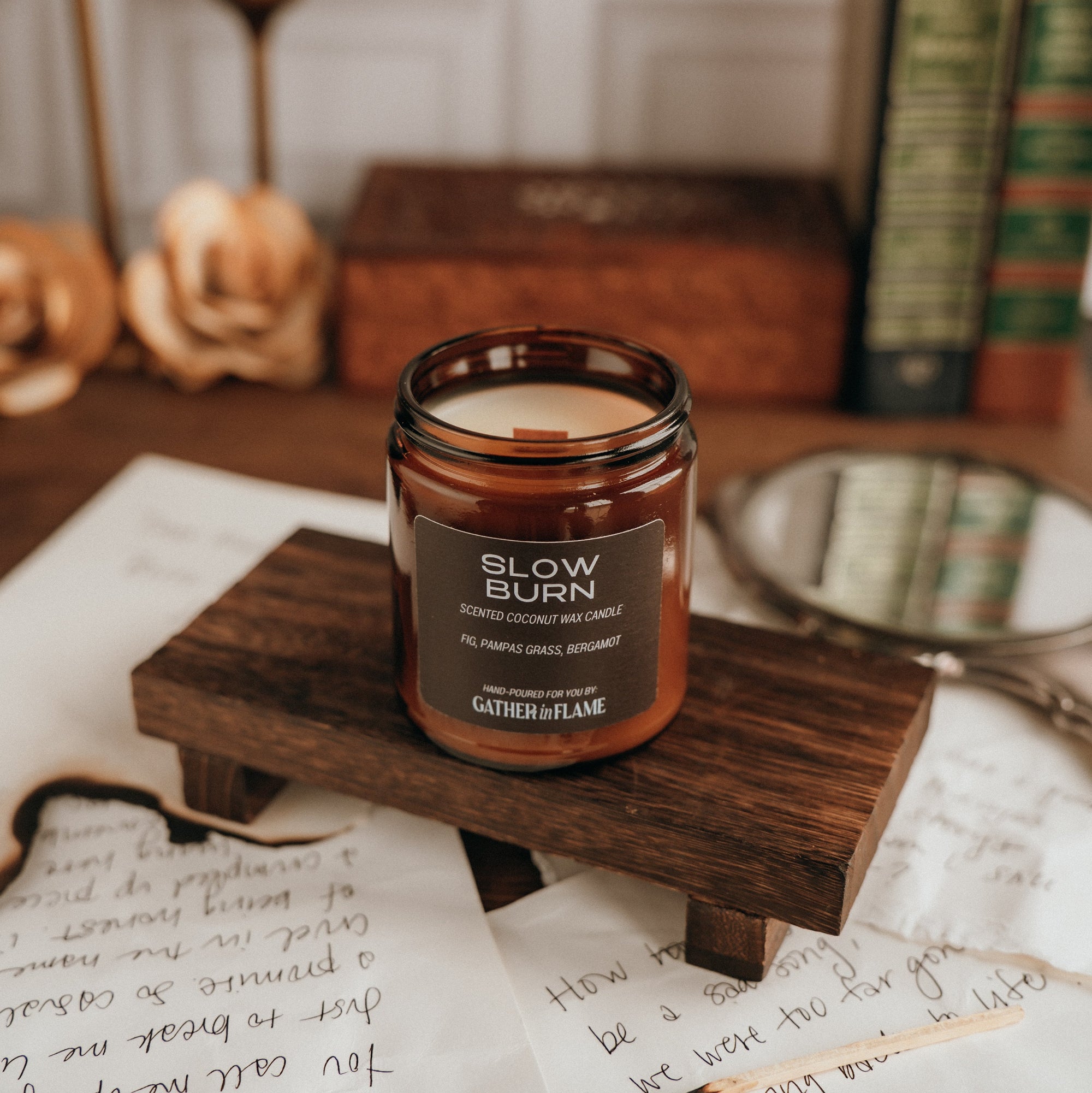 Candle labeled 'Slow Burn' on a wooden stand with books and writing utensils in the background.