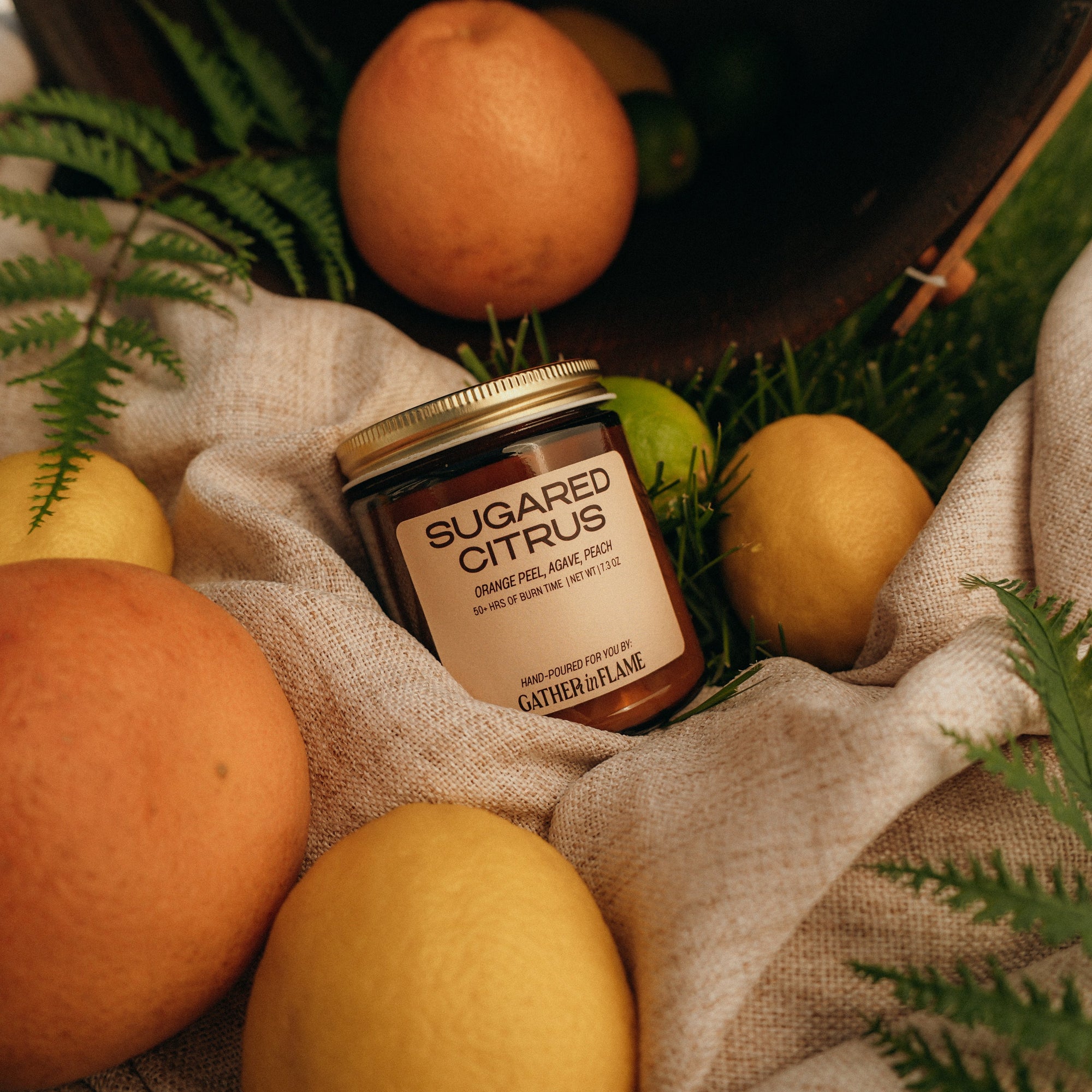 Jar of 'Sugared Citrus' candle surrounded by fruits and foliage on a textured fabric background