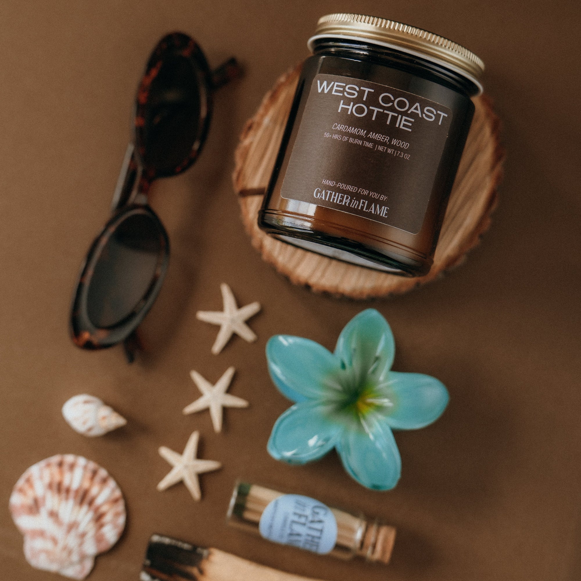 Candle labeled 'West Coast Hottie' surrounded by beach-themed items on a brown background
