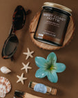 Candle labeled 'West Coast Hottie' surrounded by beach-themed items on a brown background