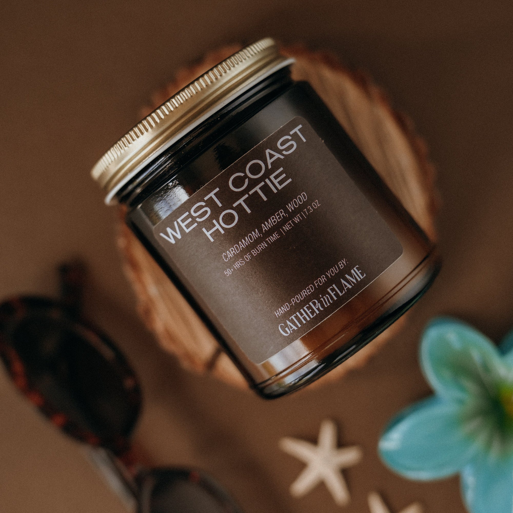 Candle jar labeled 'West Coast Hottie' by Gathered Flame on a brown background with starfish and a flower.