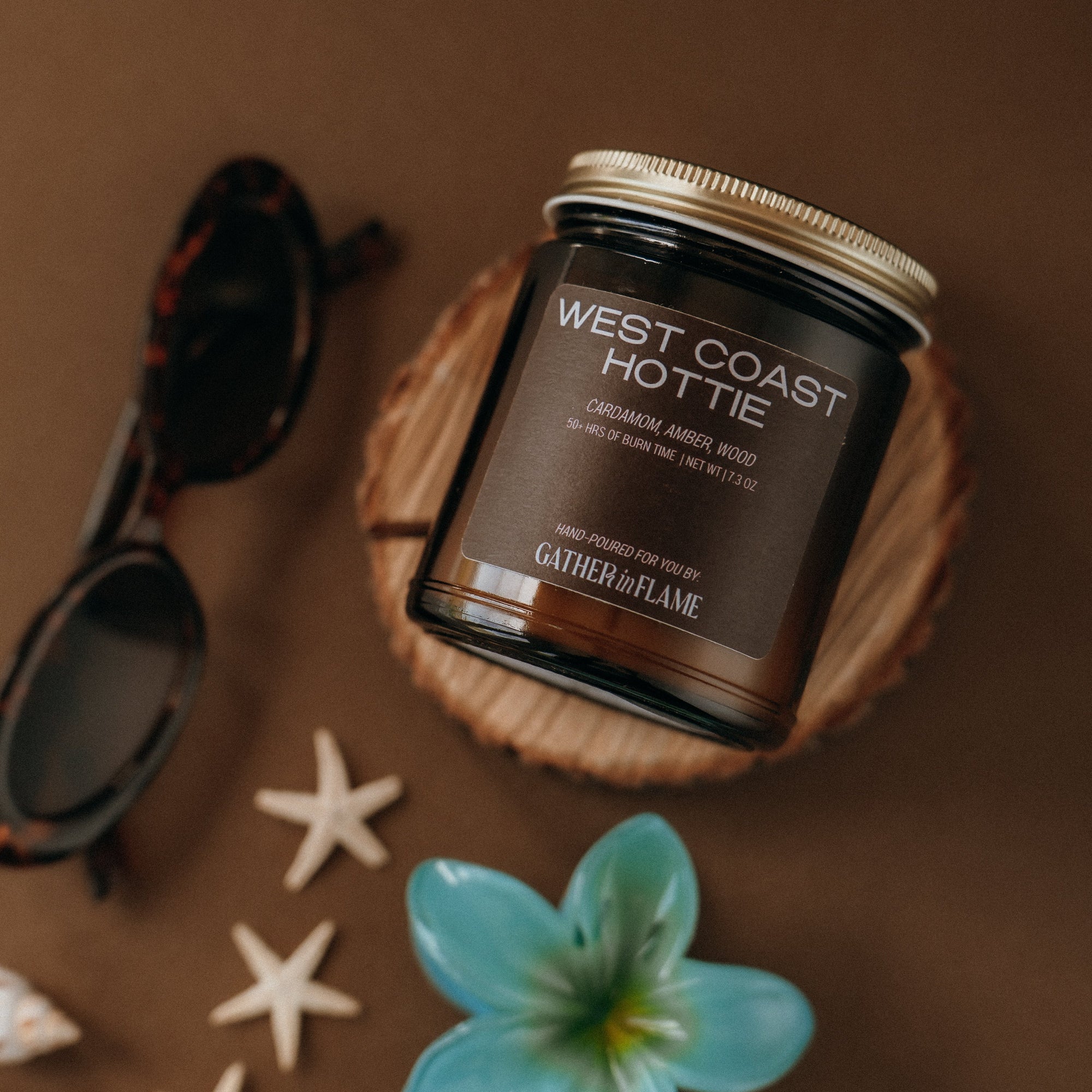 Jar of 'West Coast Hottie' candle on a wooden coaster with decorative items on a brown background