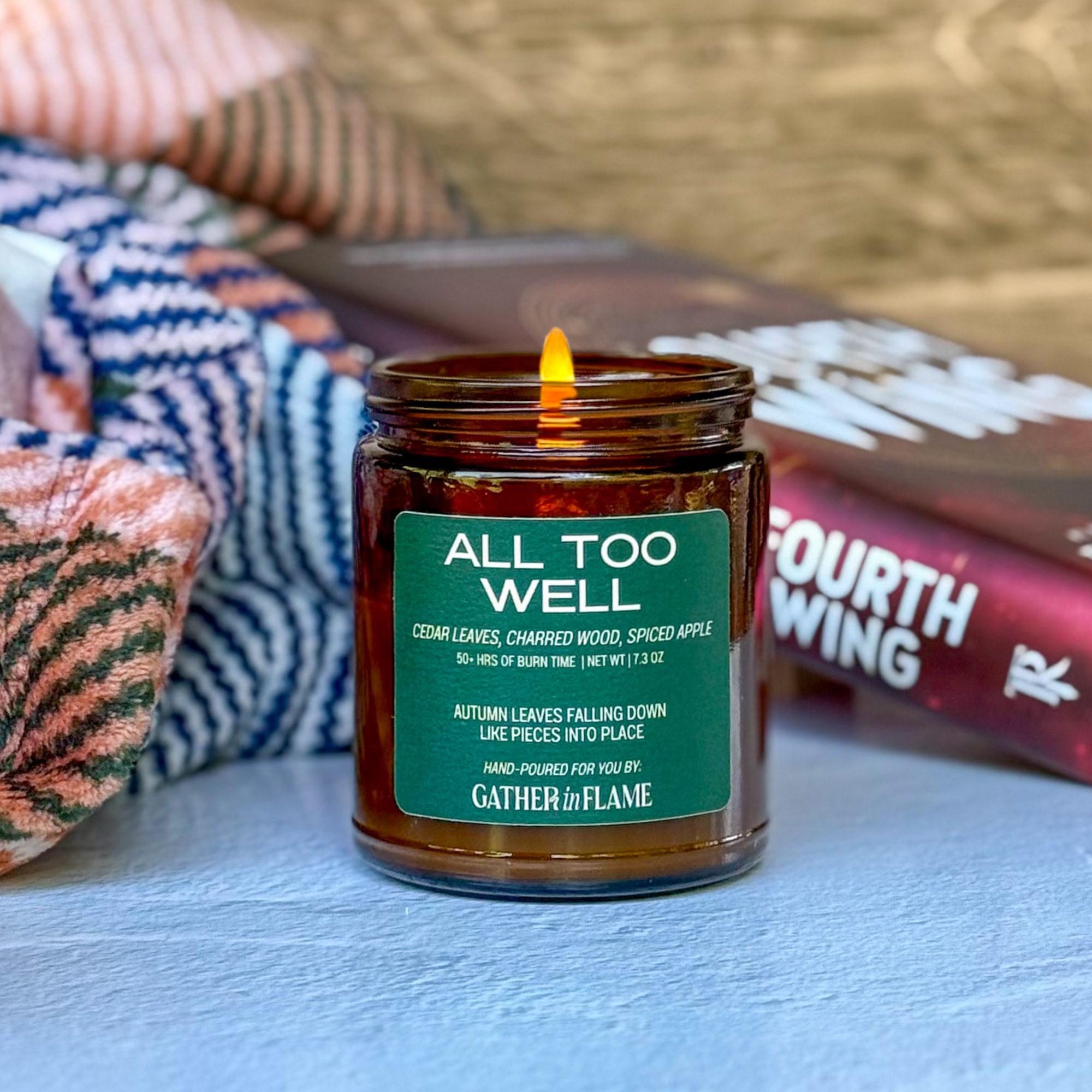 Candle in a amber jar with a green label on a surface with books and a blanket.