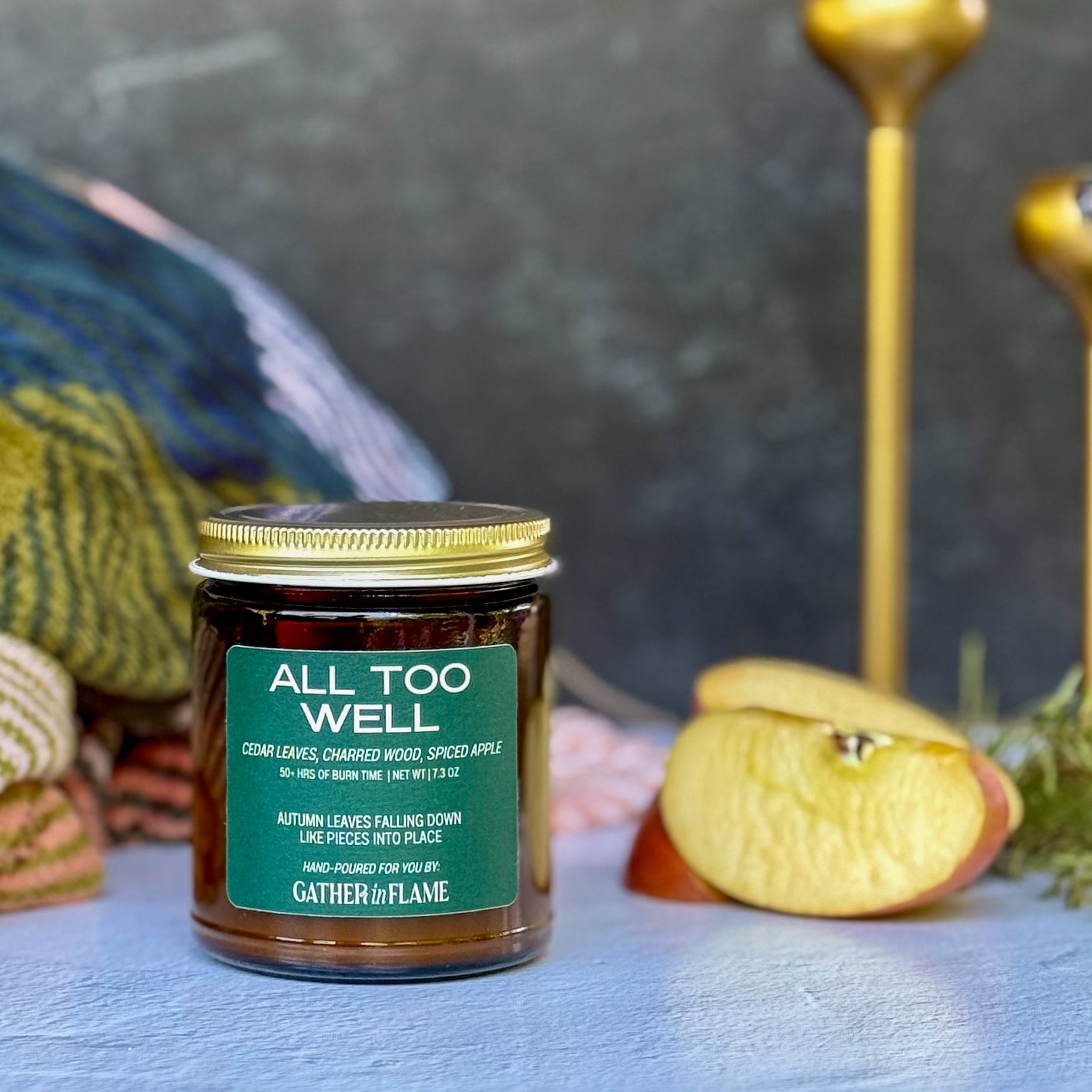 Jar of 'All Too Well' candle with a green label on a textured surface with apples and decorative elements.
