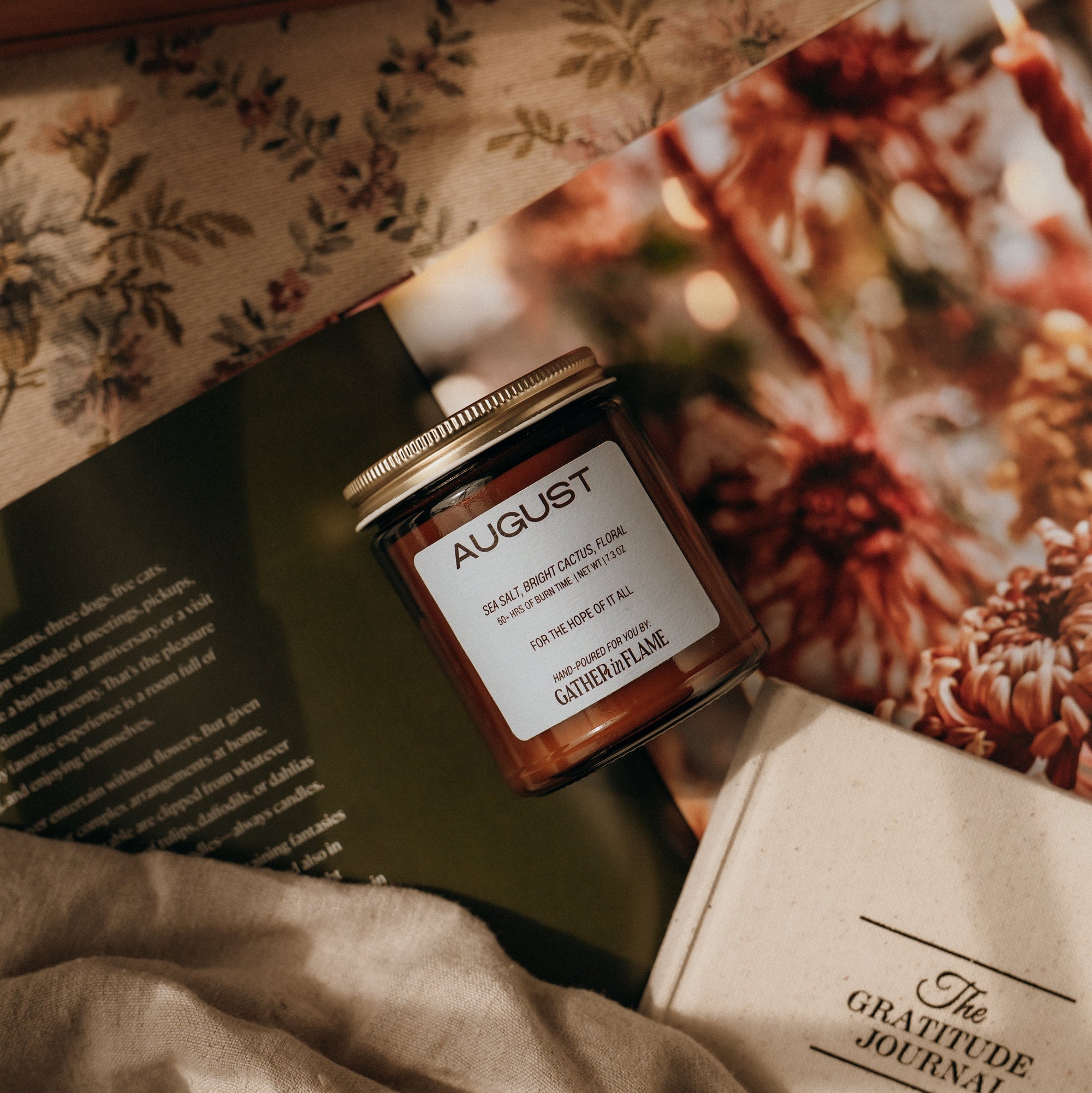 Jar of August candle on a surface with a Gratitude Journal and floral photo in the background