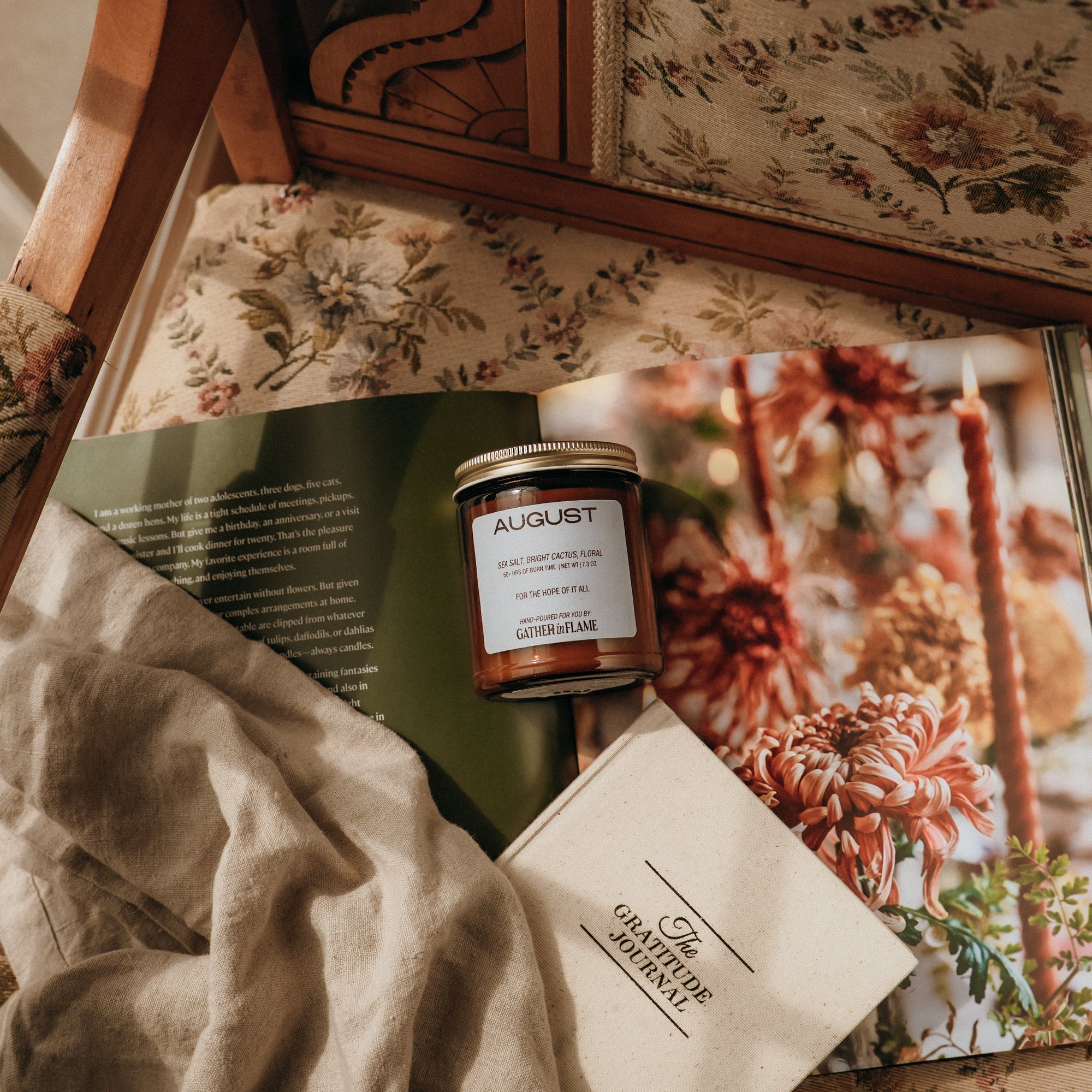 Candle with a label on top of a magazine featuring floral images, all on a floral-patterned surface.