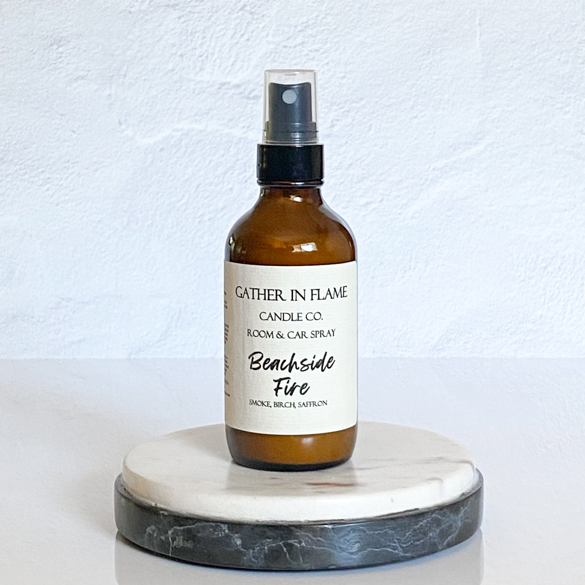 A brown amber spray bottle labeled 'Gather In Flame Candle Co. Beachside Fire Room & Car Spray' placed on a white marble surface.
