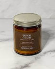 Amber glass jar candle labeled Book Club close up with a white background.