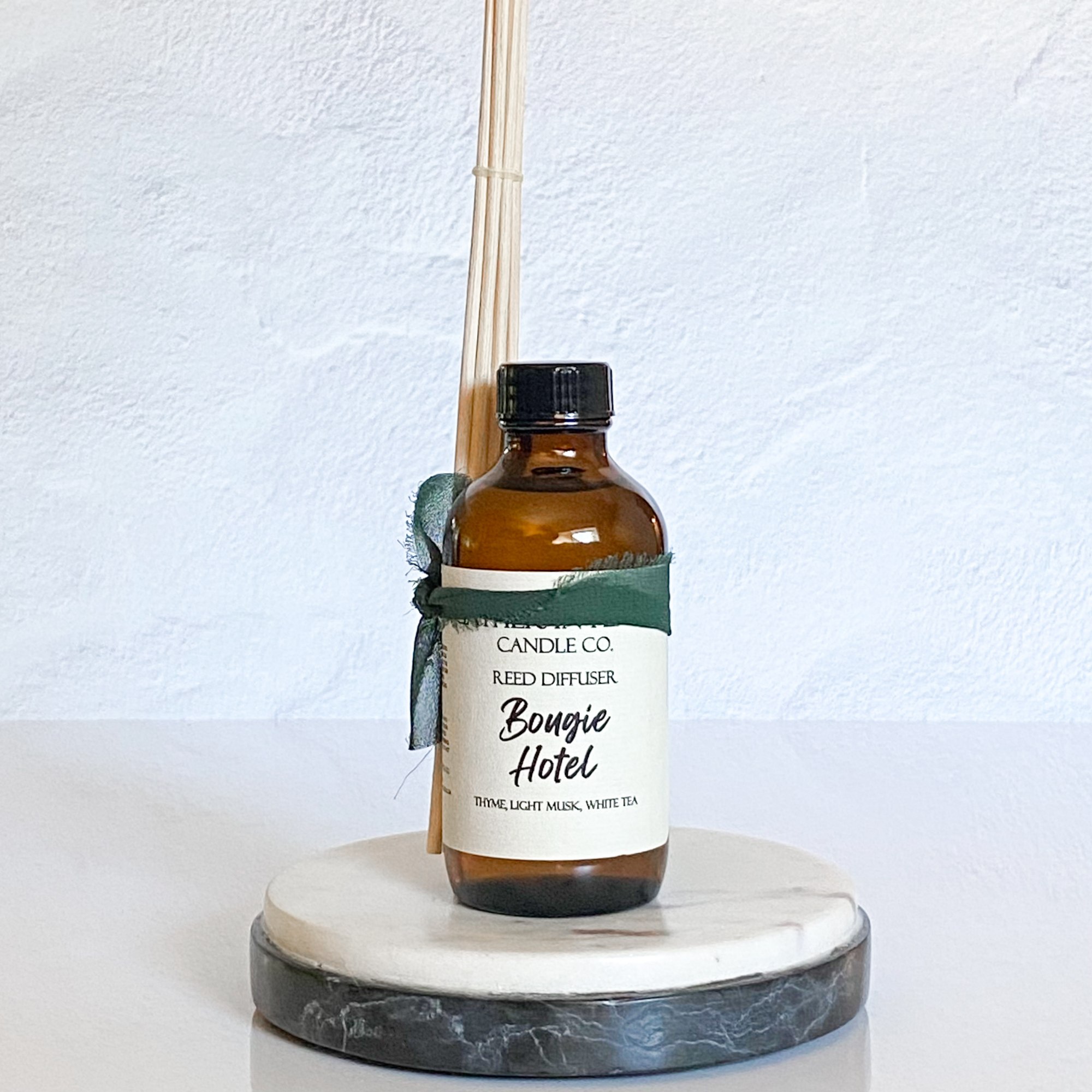 A brown glass reed diffuser bottle with a label reading "Bougie Hotel", placed on a white marble round tray, with reeds inserted into the bottle for diffusing the fragrance.