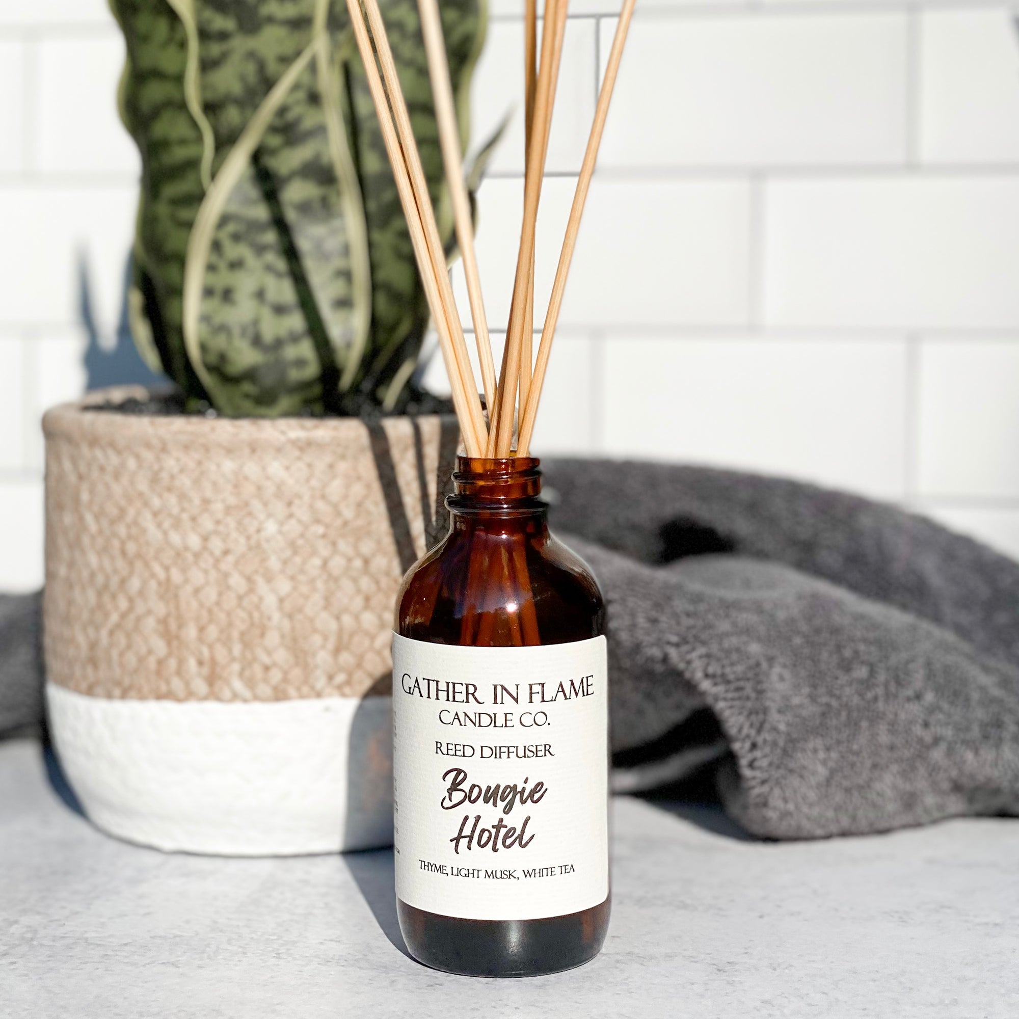 A brown glass reed diffuser bottle with a label reading "Bougie Hotel", placed grey surface, surrounded by plant and towels with white tile background.