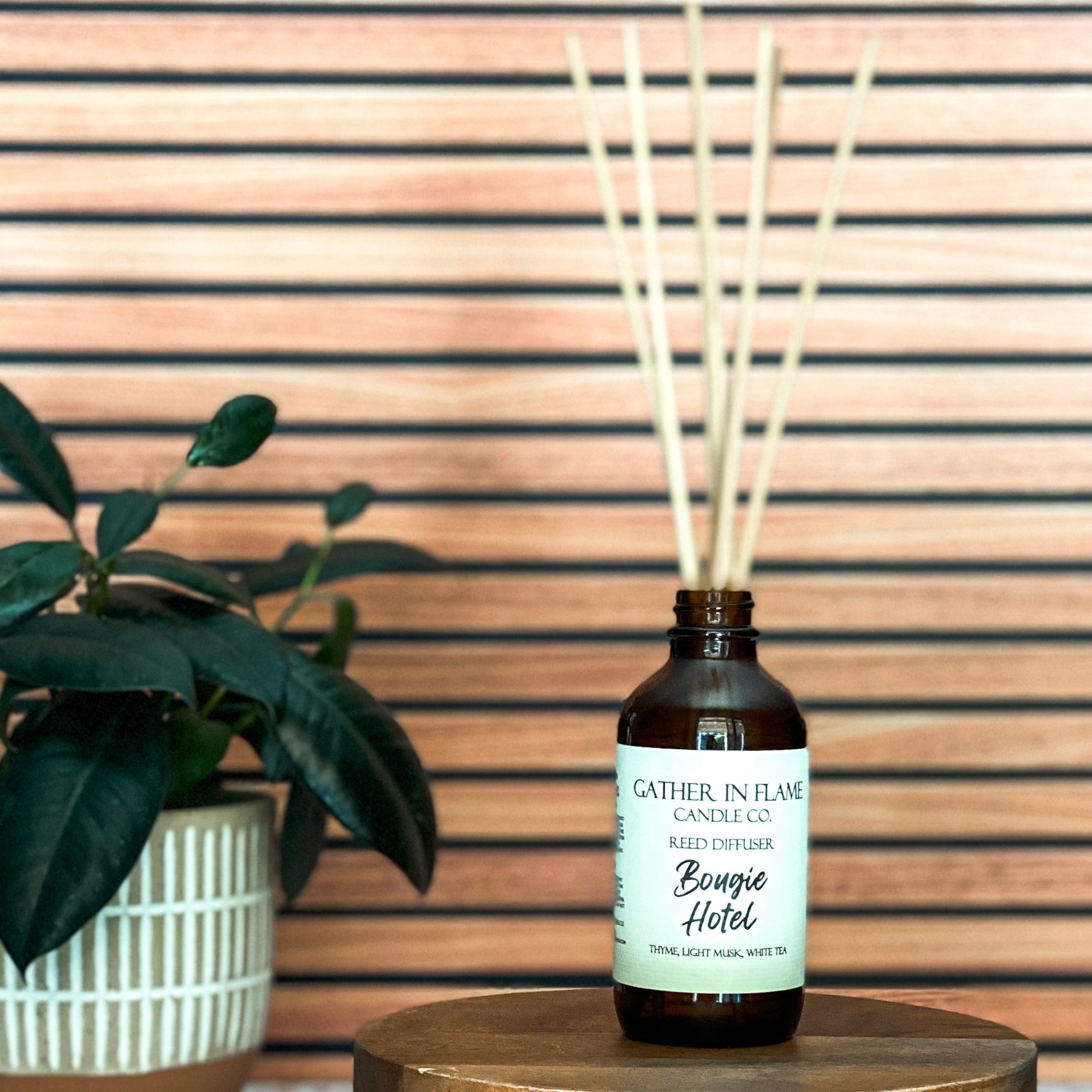 A brown glass reed diffuser bottle with a label reading "Bougie Hotel", placed on a wood surface, with plant and wood slat background.