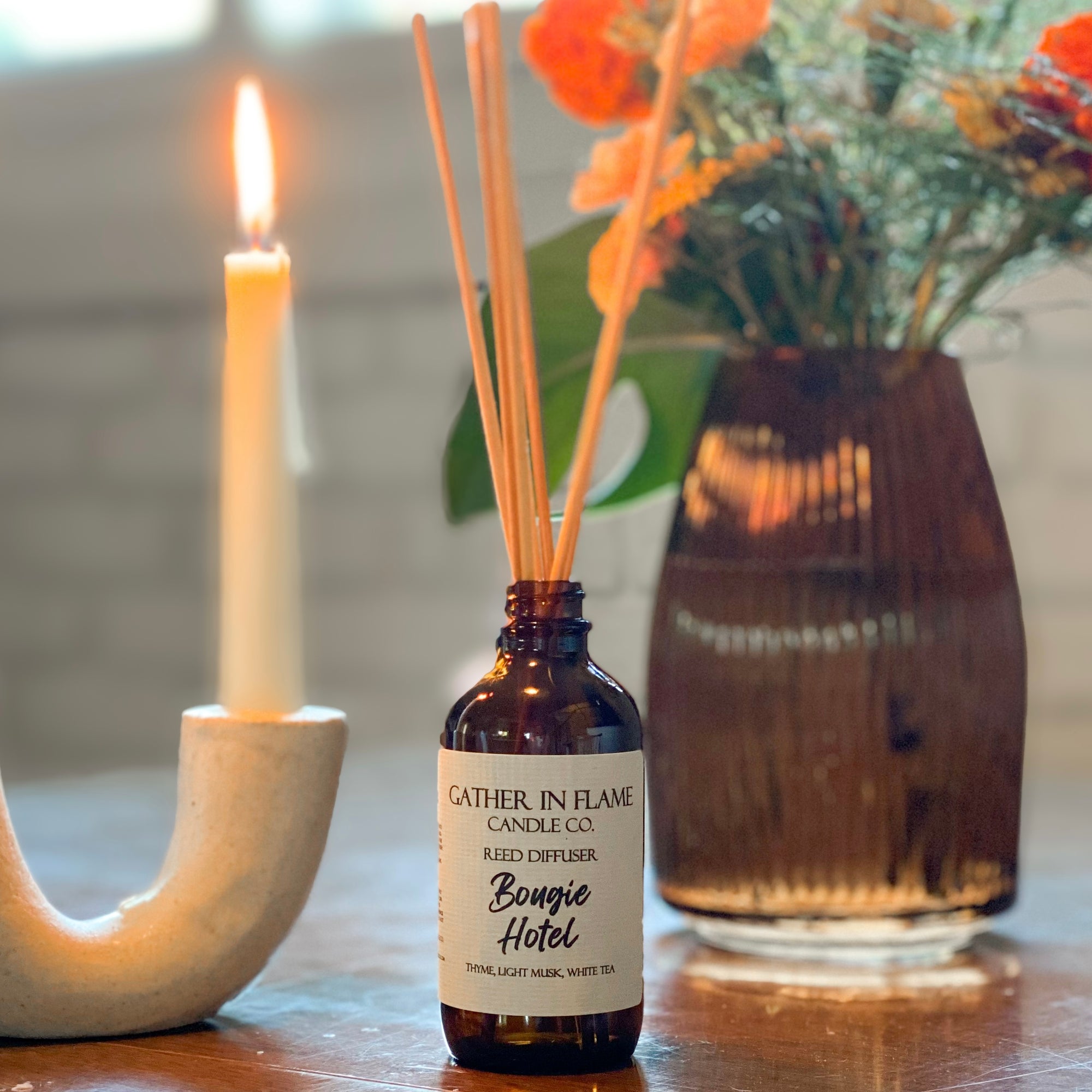 A brown glass reed diffuser bottle with a label reading "Bougie Hotel", placed on table with candle and flowers in a vase in the background.