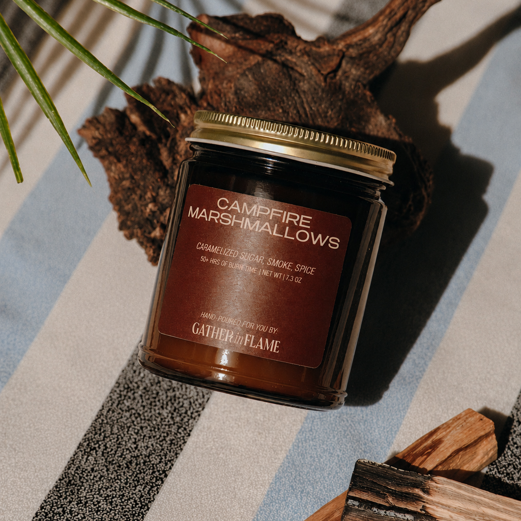 Jar of 'Campfire Marshmallows' candle on a striped surface with wooden elements and a plant.