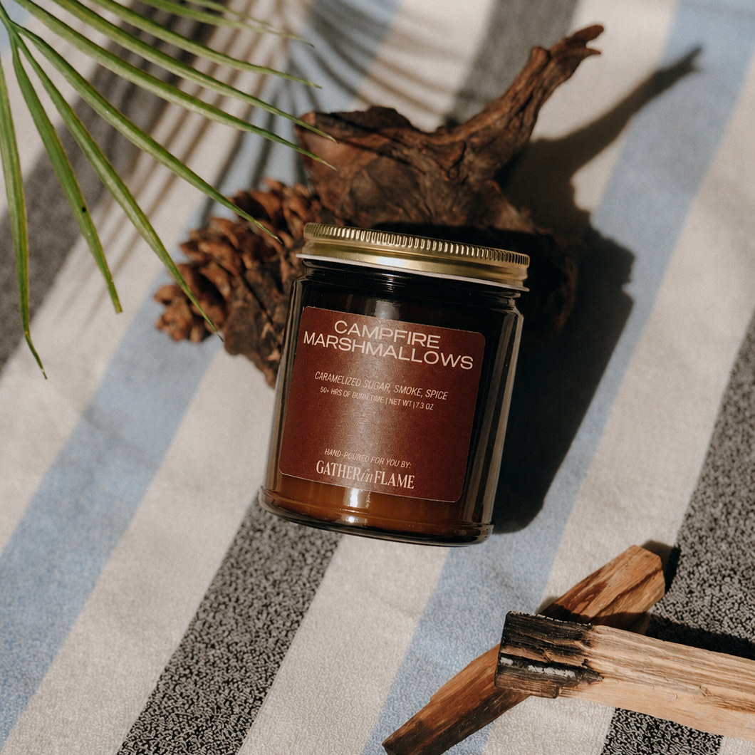 Jar of 'Campfire Marshmallows' candle on a striped fabric background with natural elements.