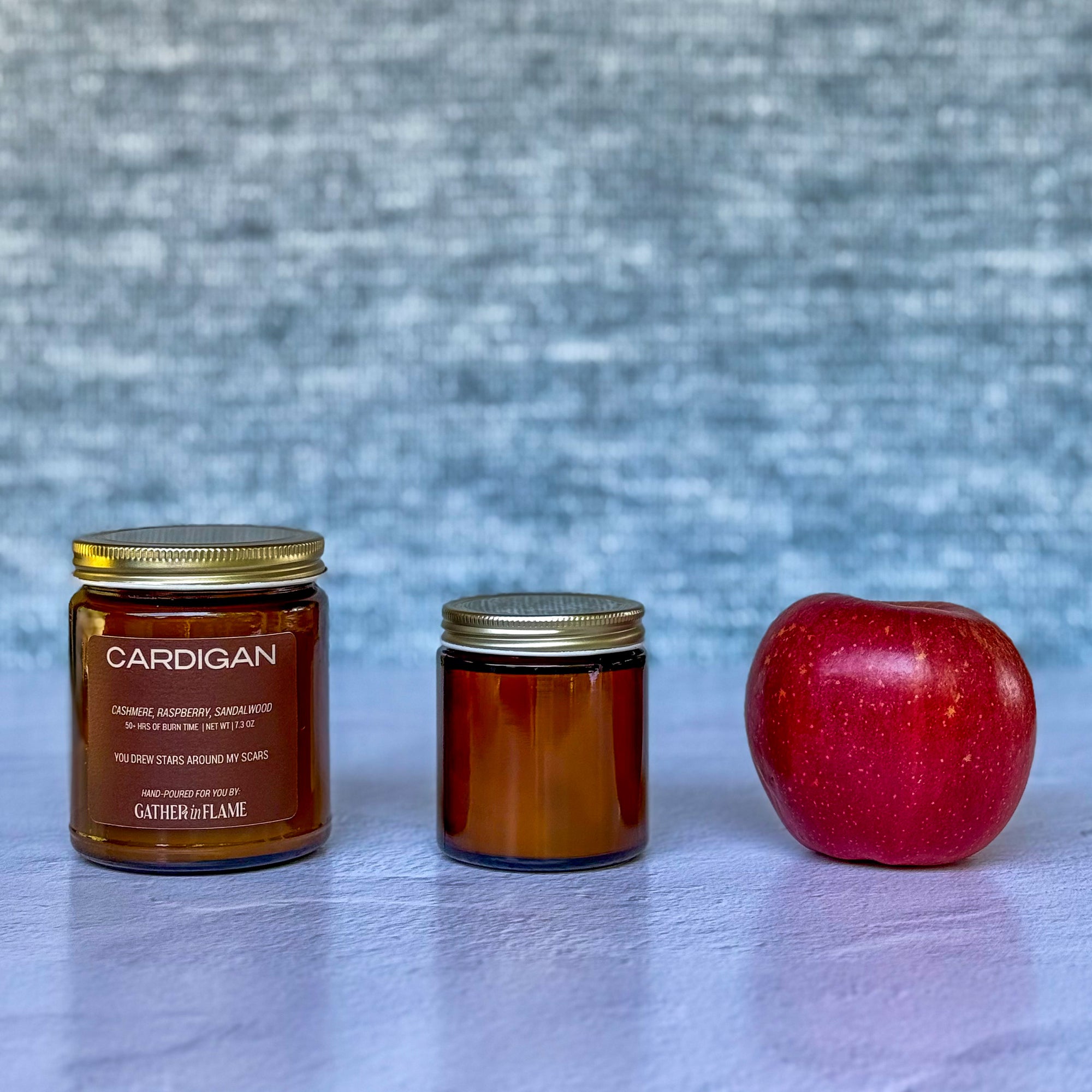 Two candles labeled Cardigan and a red apple on a textured surface with a gray background