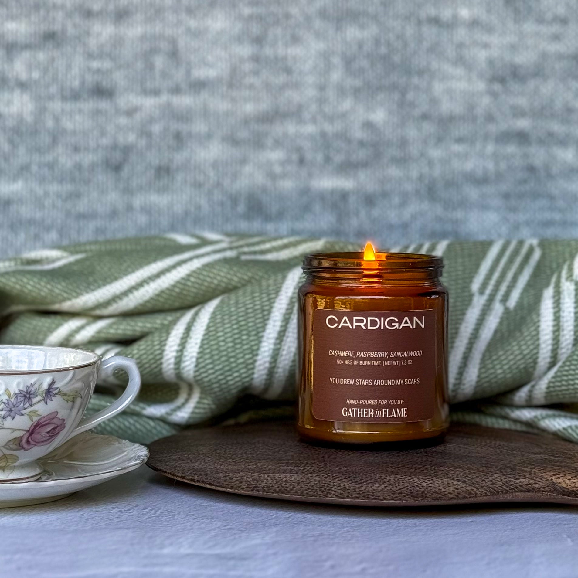 Candle in a brown jar labeled 'Cardigan' with a teacup and green blanket in the background.