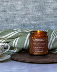 Candle in a brown jar labeled 'Cardigan' with a teacup and green blanket in the background.