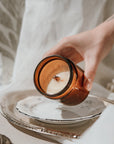 Hand holding a small amber glass jar with a candle on a white surface