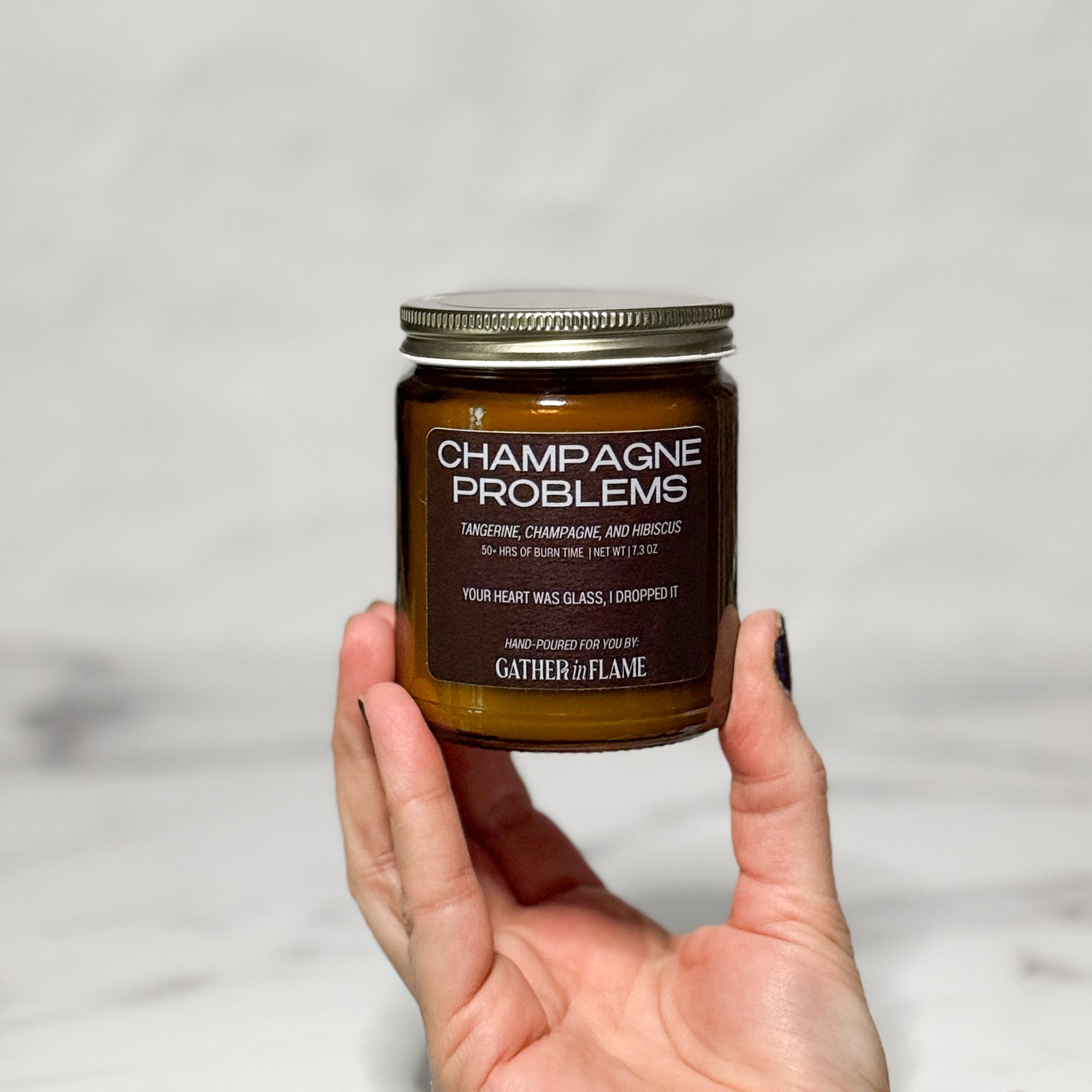 Candle jar labeled 'Champagne Problems' held by a hand against a light gray background