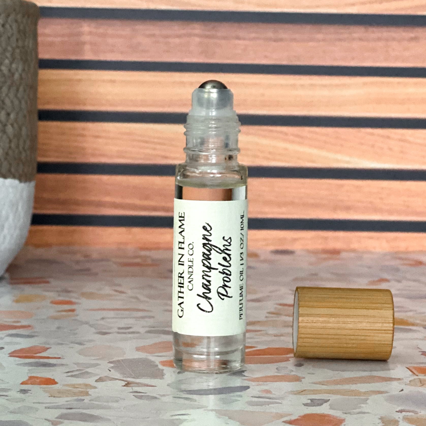 A perfume oil roller with a clear glass body and bamboo cap sitting beside it, labeled 'Champagne Problems', resting on a terrazzo surface with wood background.