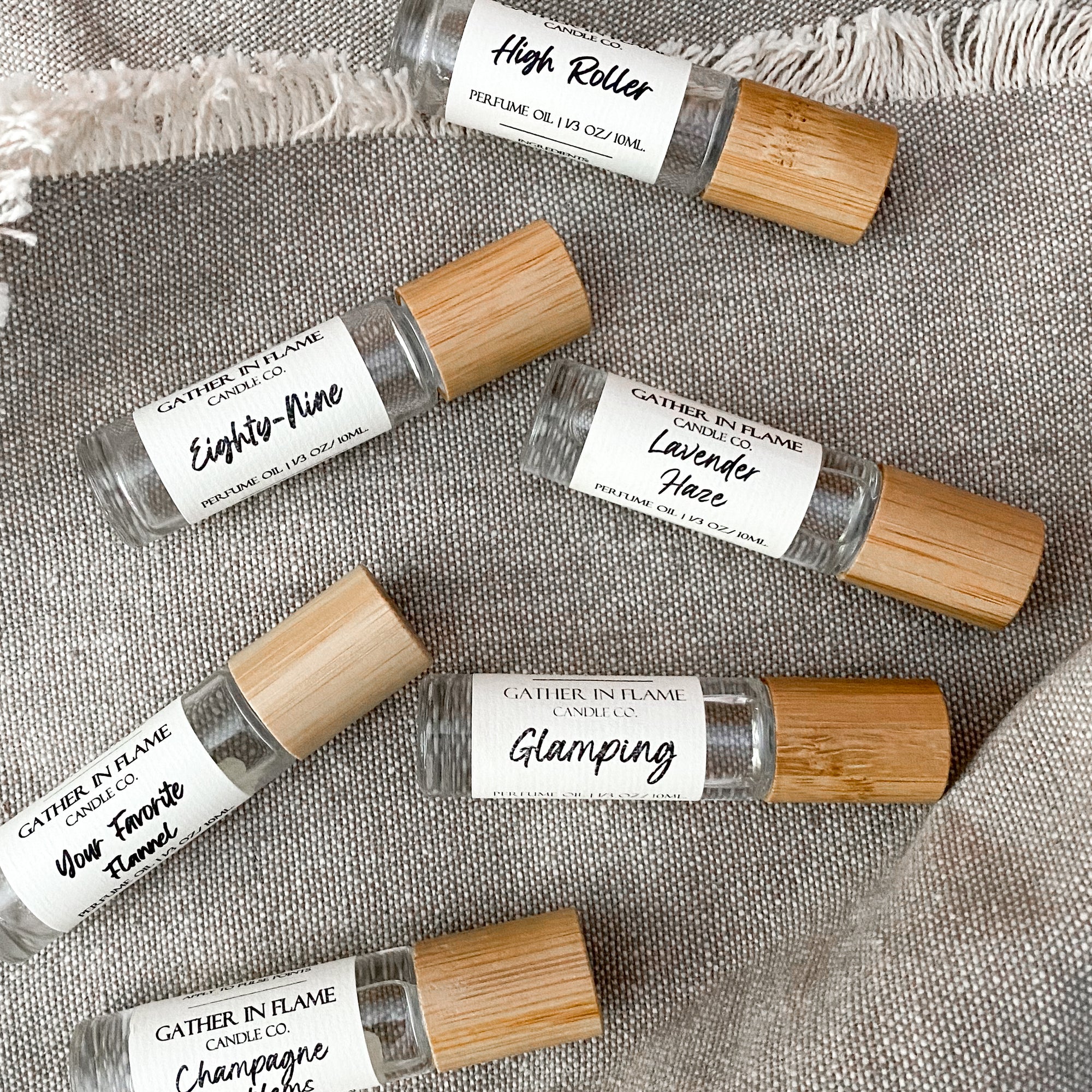 Flat lay of six perfume oil rollers with clear glass body and bamboo cap in a diagonal line resting on a tan cloth surface.