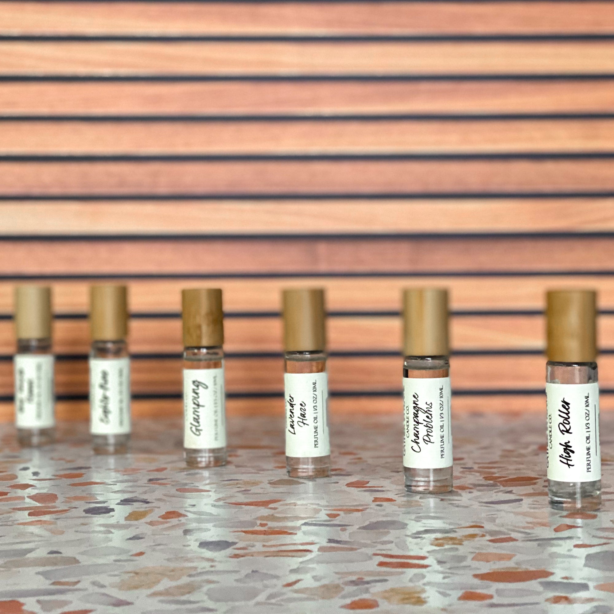 Six perfume oil roller with  clear glass body and bamboo cap in a diagonal line resting on a terrazzo surface with wood background.