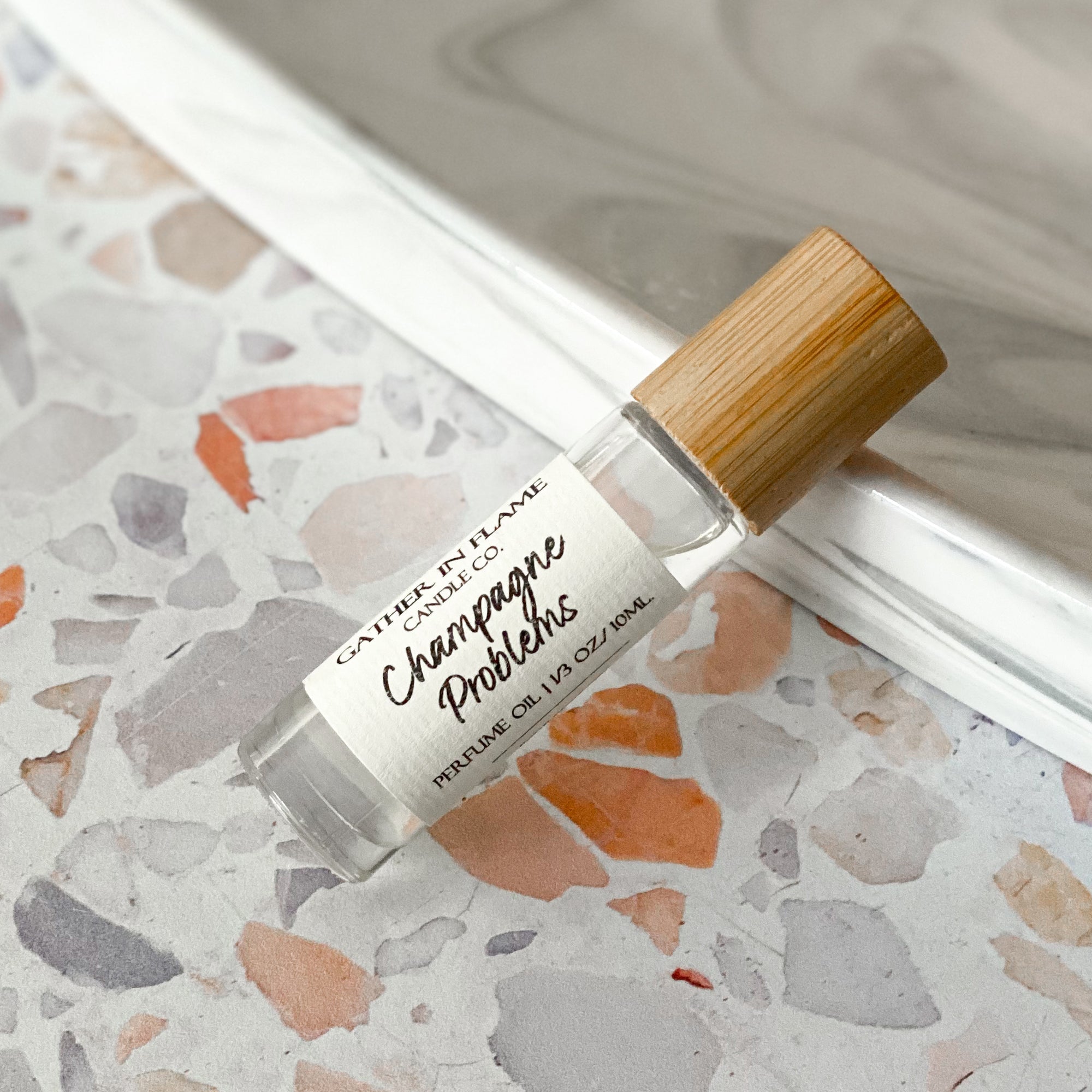 A perfume oil roller with a clear glass body and bamboo cap, labeled 'Champagne Problems', resting on a terrazzo surface.