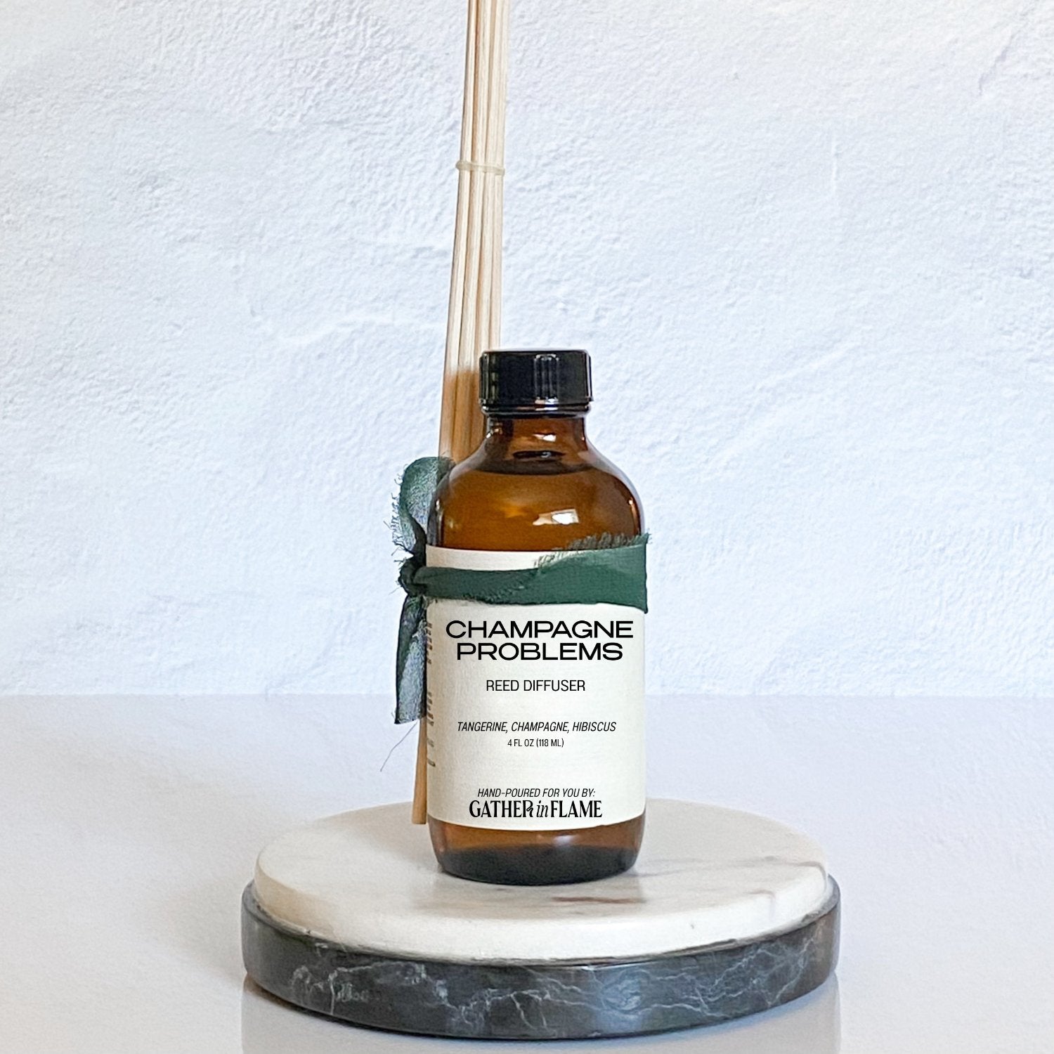 Amber glass reed diffuser bottle labeled Champagne Problems wrapped in a green ribbon, placed on a marble coaster with a white background.