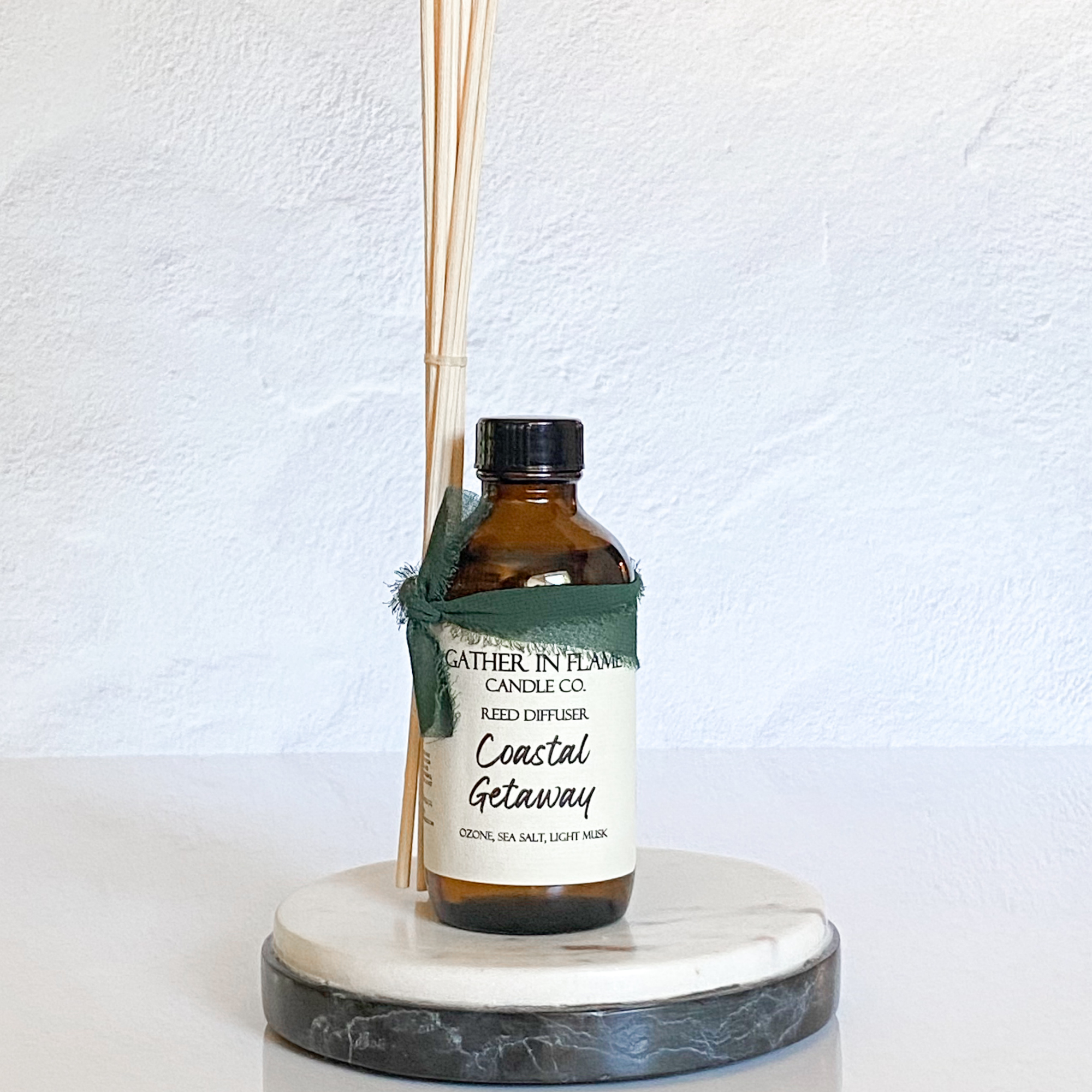 A Reed diffuser with amber glass bottle placed on a round white marble coaster. The label on the bottle reads 'Coastal Getaway'.