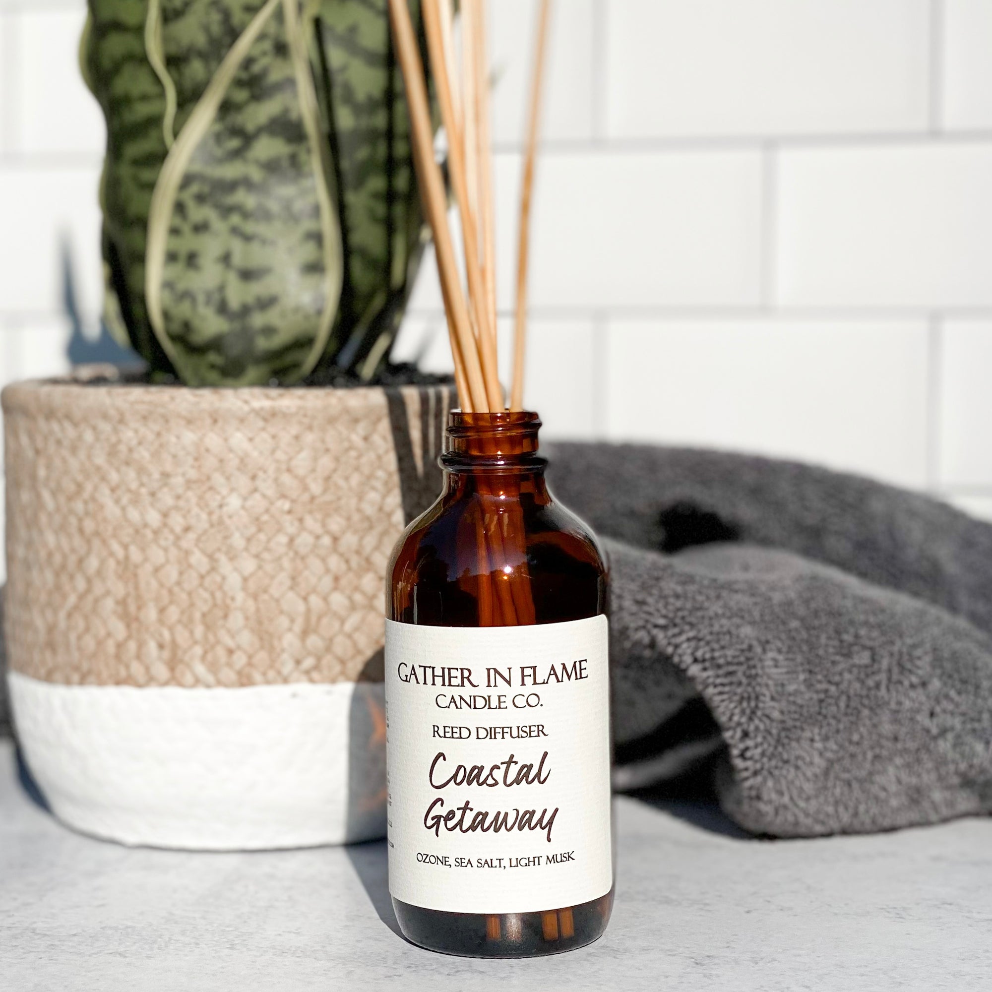 A brown glass reed diffuser bottle with a label reading "Coastal Getaway", placed grey surface, surrounded by plant and towels with white tile background.