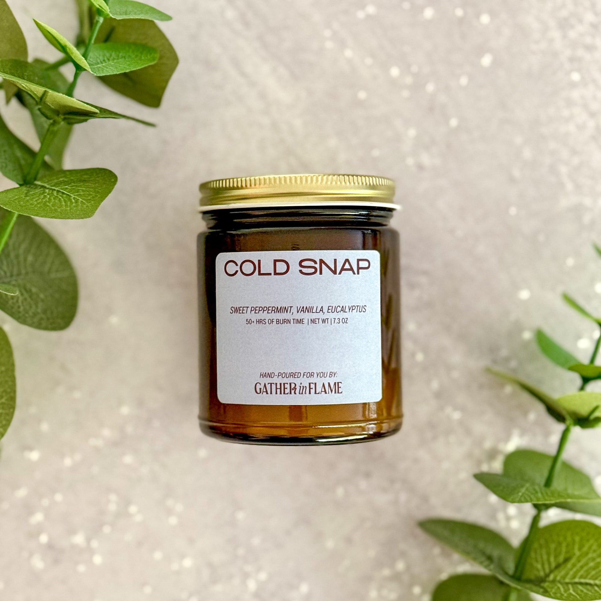 Candle jar labeled 'Cold Snap' with a gold lid on a white background with green leaves.