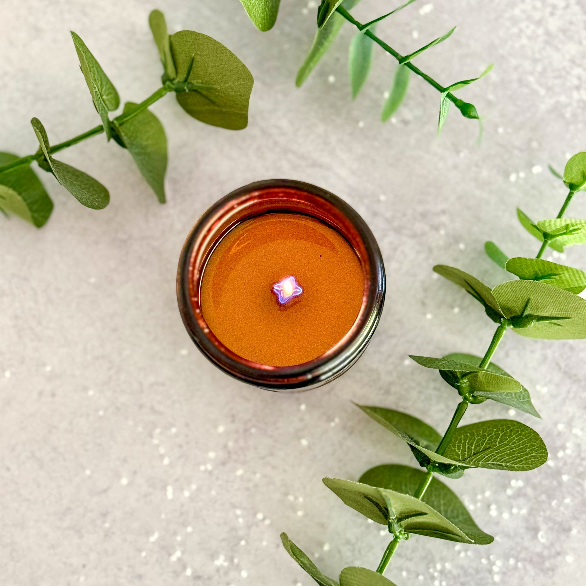 Overhead view of lit candle surface with melted wax surrounded by green leaves on a textured white background