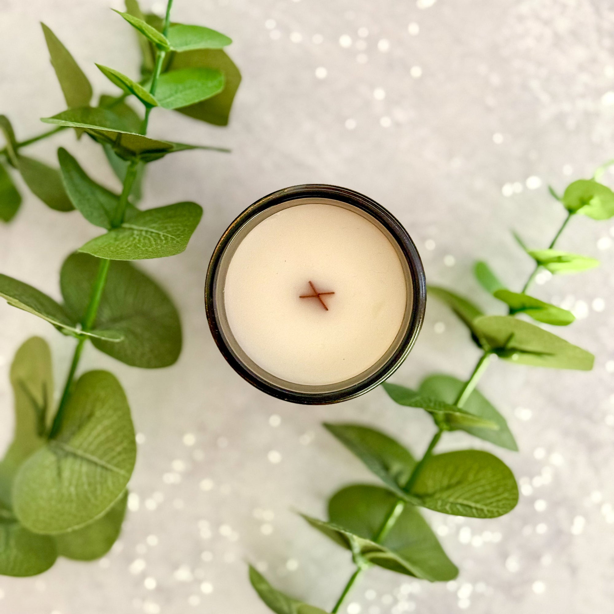 Overhead view of candle surface with wood wick surrounded by green leaves on a textured white background