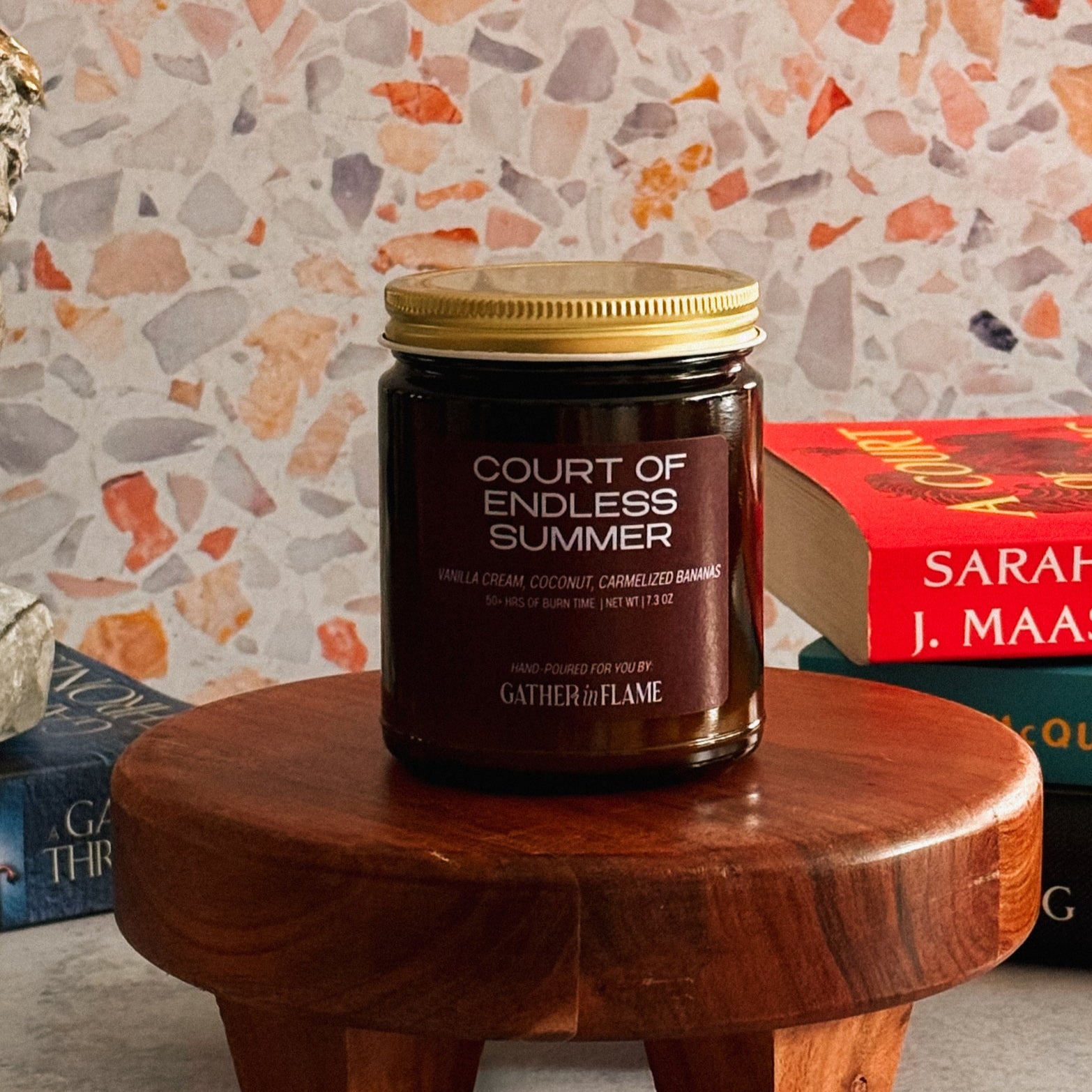 Candle jar labeled 'Court of Endless Summer' on a wooden stool with a colorful wall and books in the background