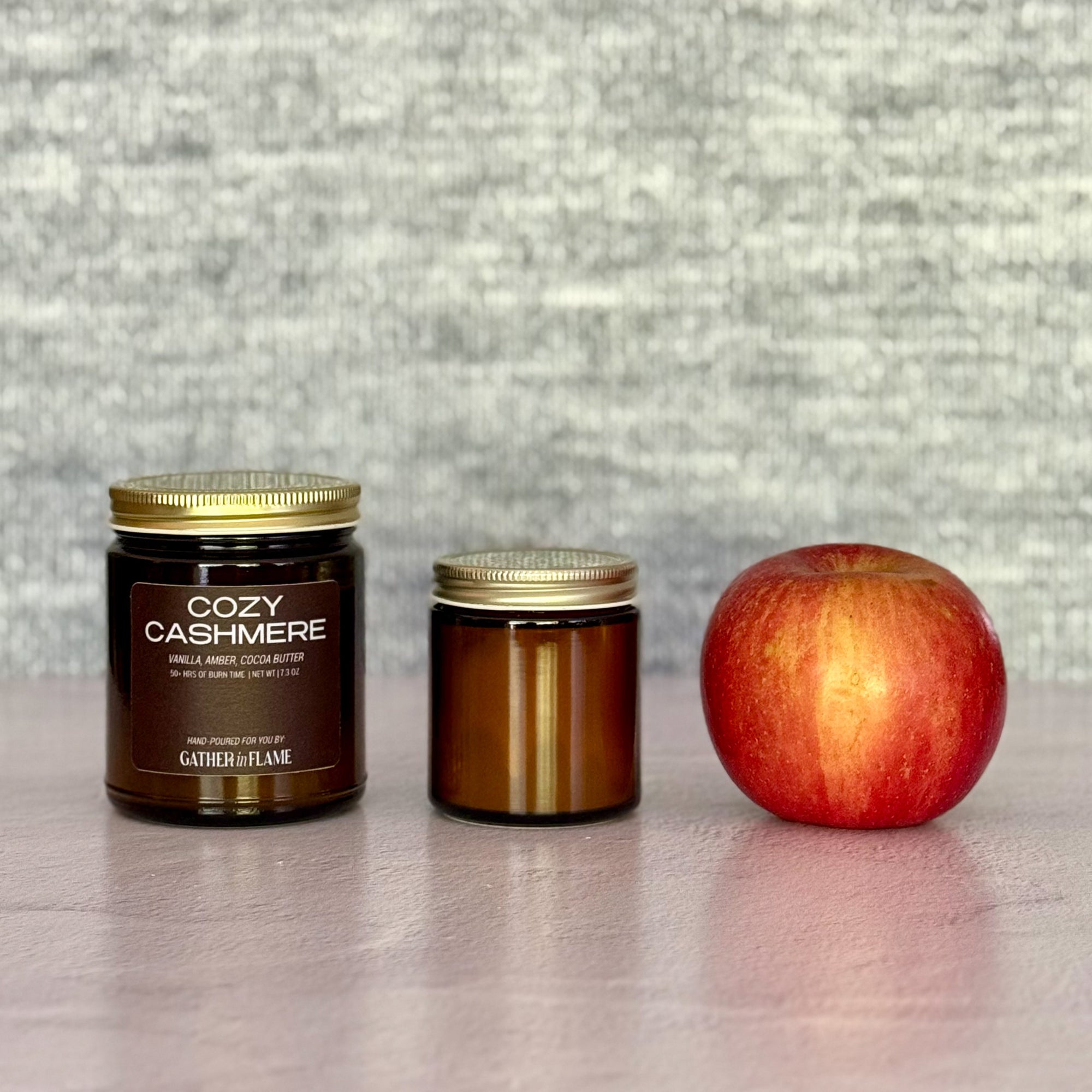 Two candles  of different sizes labeled 'Cozy Cashmere' next to an apple on a textured surface.