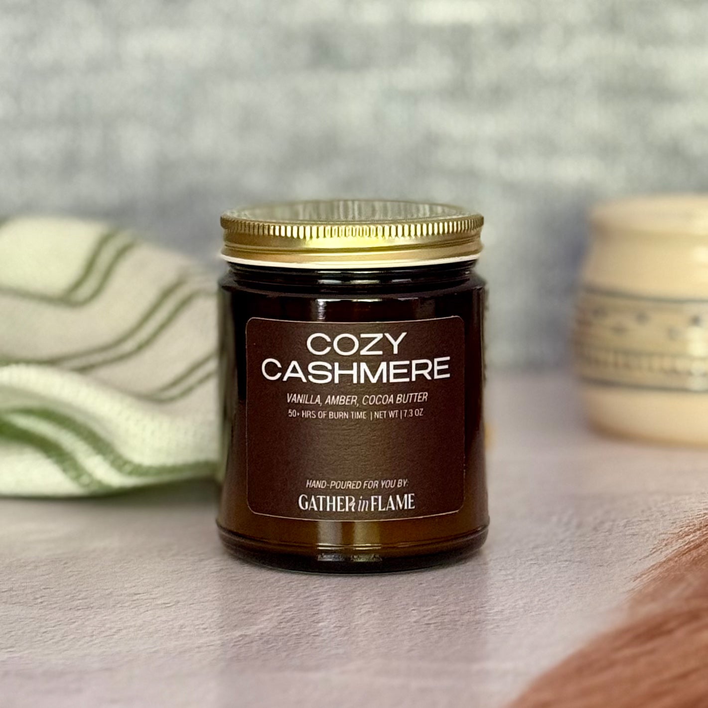 Jar of 'Cozy Cashmere' candle with gold lid on a surface with dried pampas grass, a soft blanket and mug in the background