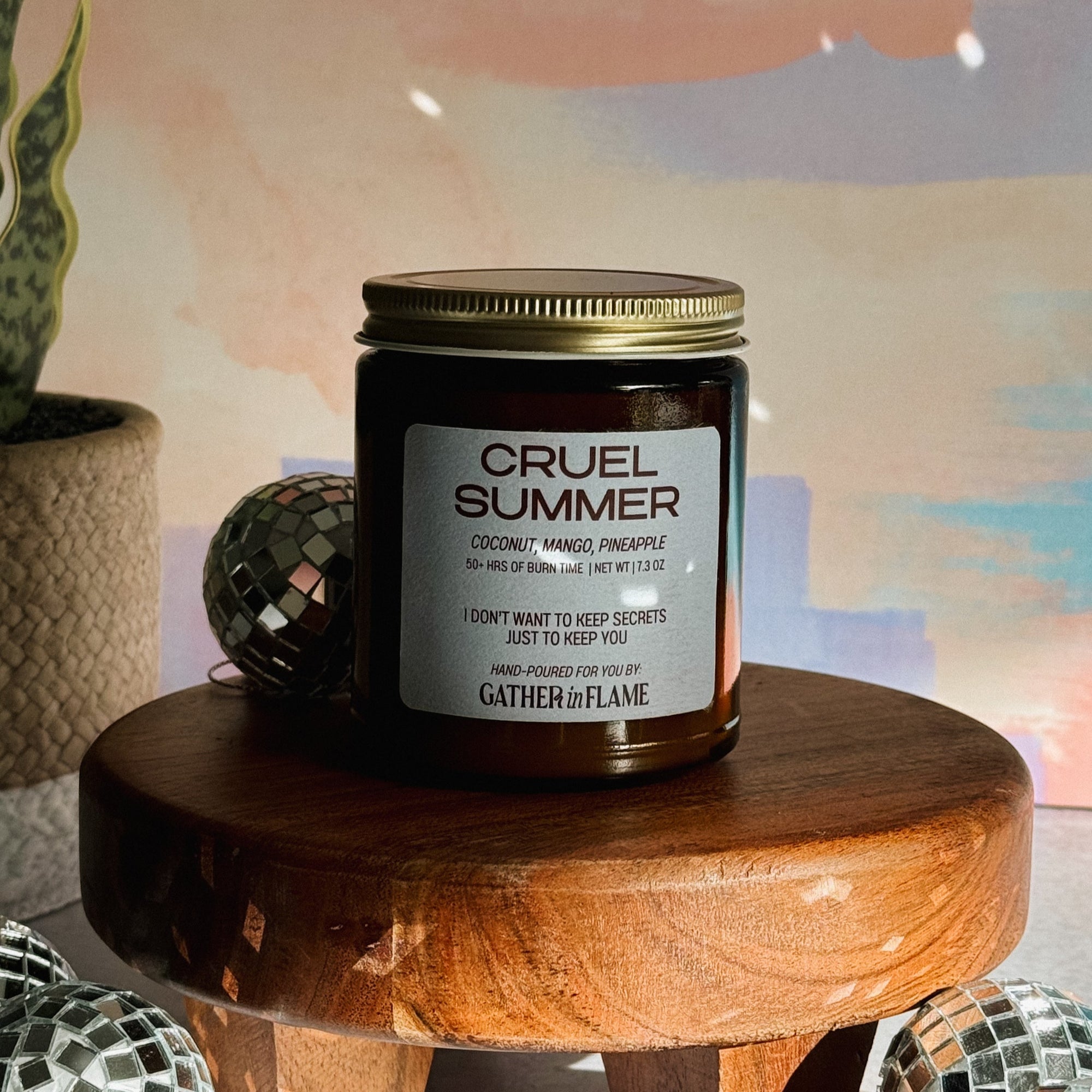 Candle labeled 'Cruel Summer' on a wooden stool with decorative items in the background