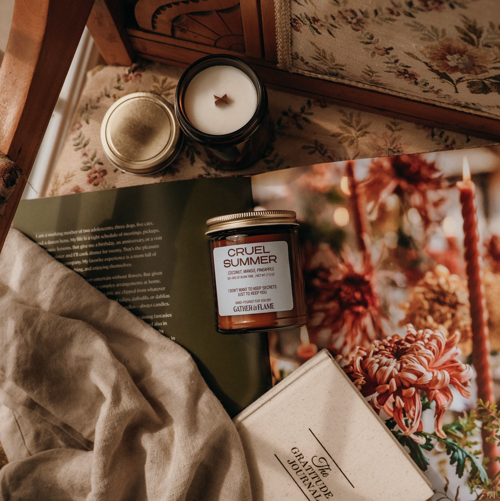 Jar of 'Cruel Summer' candle on a textured surface with a floral book and fabric.