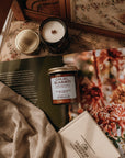 Jar of 'Cruel Summer' candle on a textured surface with a floral book and fabric.