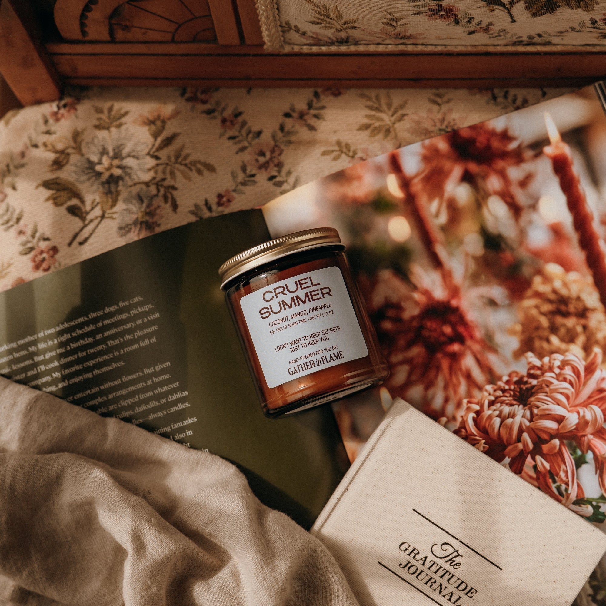 Jar of 'Cruel Summer' candle on a floral-patterned surface with a book and fabric.