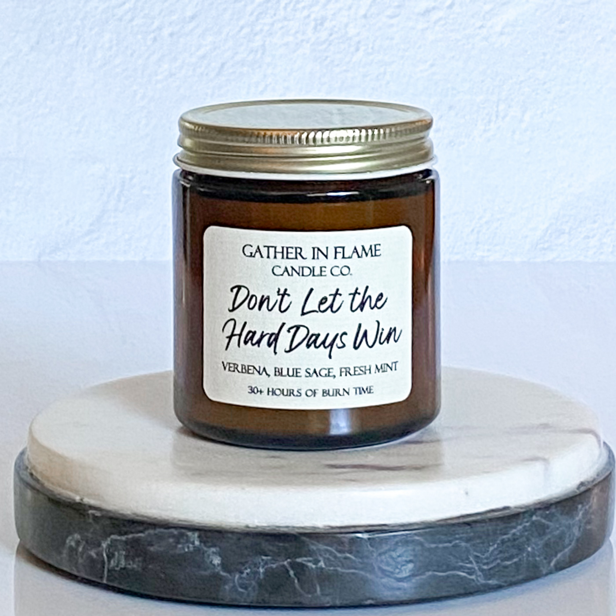 A coconut wax candle jar with a label that reads 'Don't Let the Hard Days Win', placed on a marble surface, with a white background and a gold lid on top.