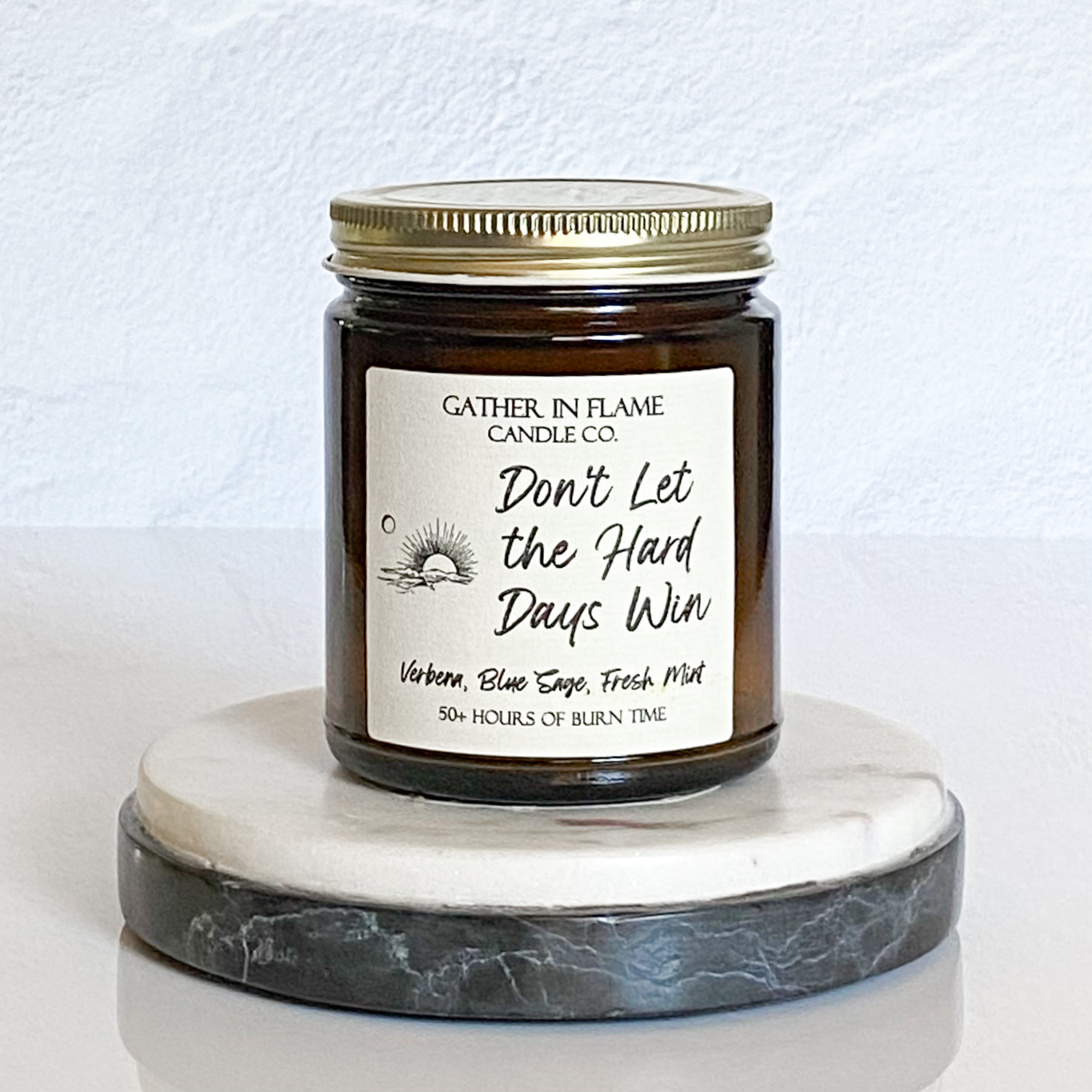 A coconut wax candle jar with a label that reads 'Don't Let the Hard Days Win', placed on a marble surface, with a white background and a gold lid on top.