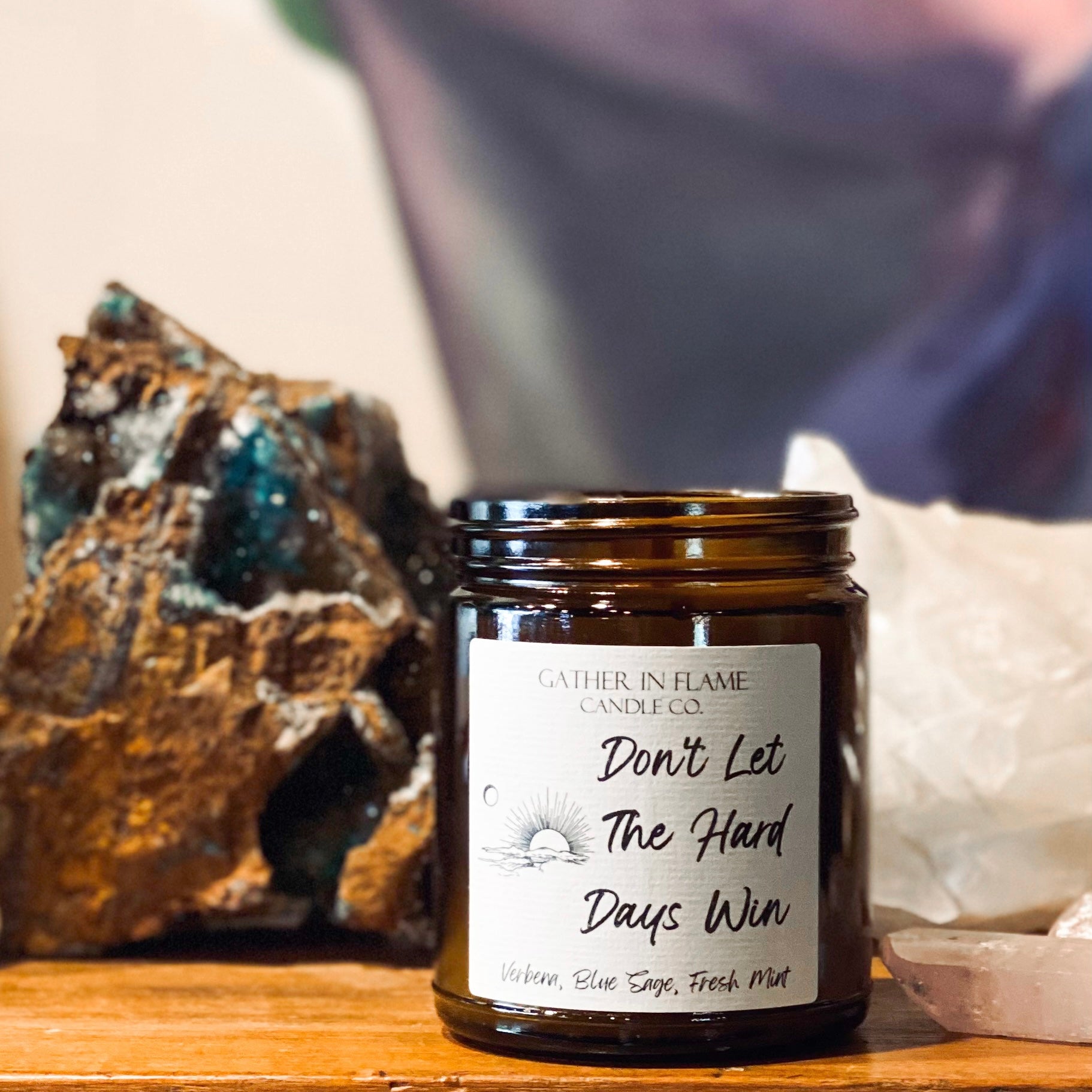 Amber jar candle labeled Don't Let the Hard Days Win on a wood surface with art in the background and large crystals.