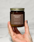 Amber jar candle labeled Dragon Rider held in a hand with a white background.