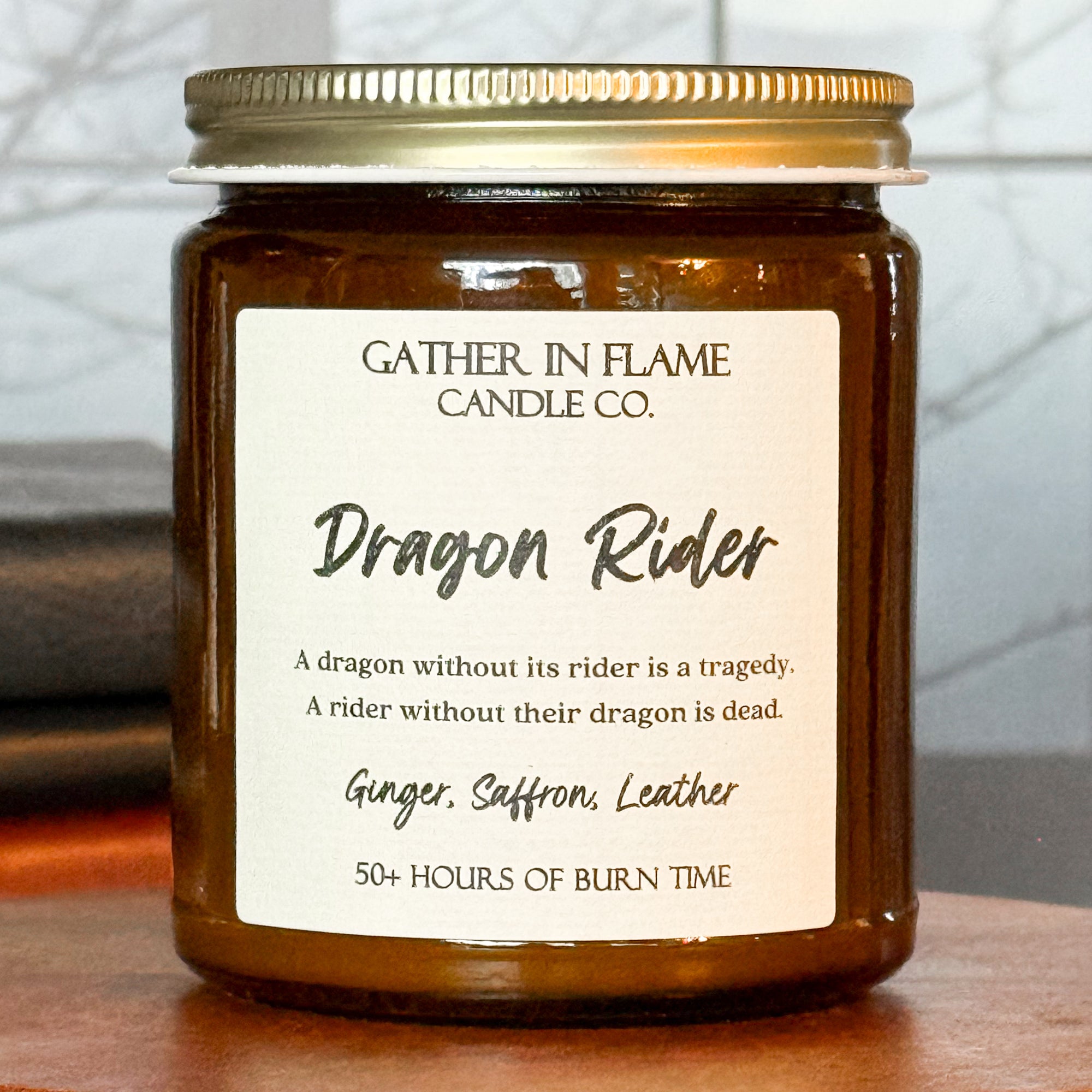 Close up of a jar candle labeled 'Dragon Rider' from Gather in Flame Candle Co.,' placed on a wooden pedestal, with books and a window in the background.
