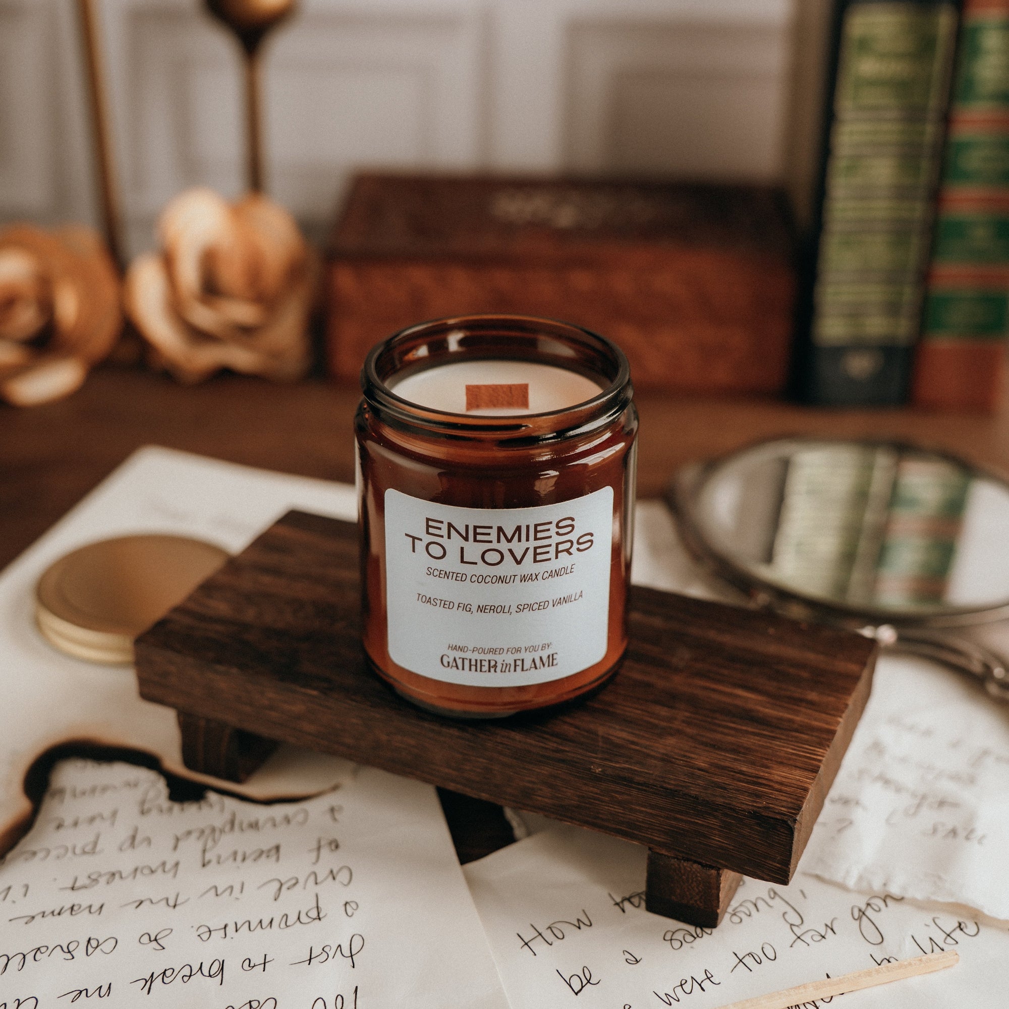 Candle labeled 'Enemies to Lovers' on a wooden stand with stationery items in the background.