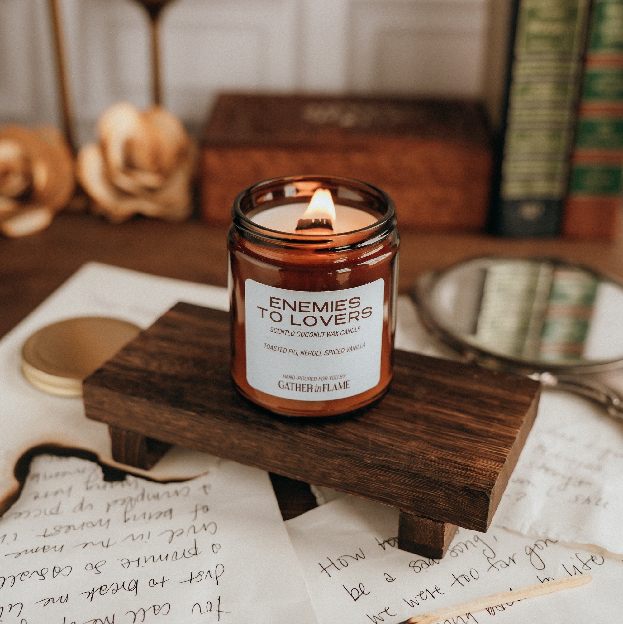 Candle in a jar labeled 'Enemies to Lovers' on a wooden stand with papers and a mirror in the background.